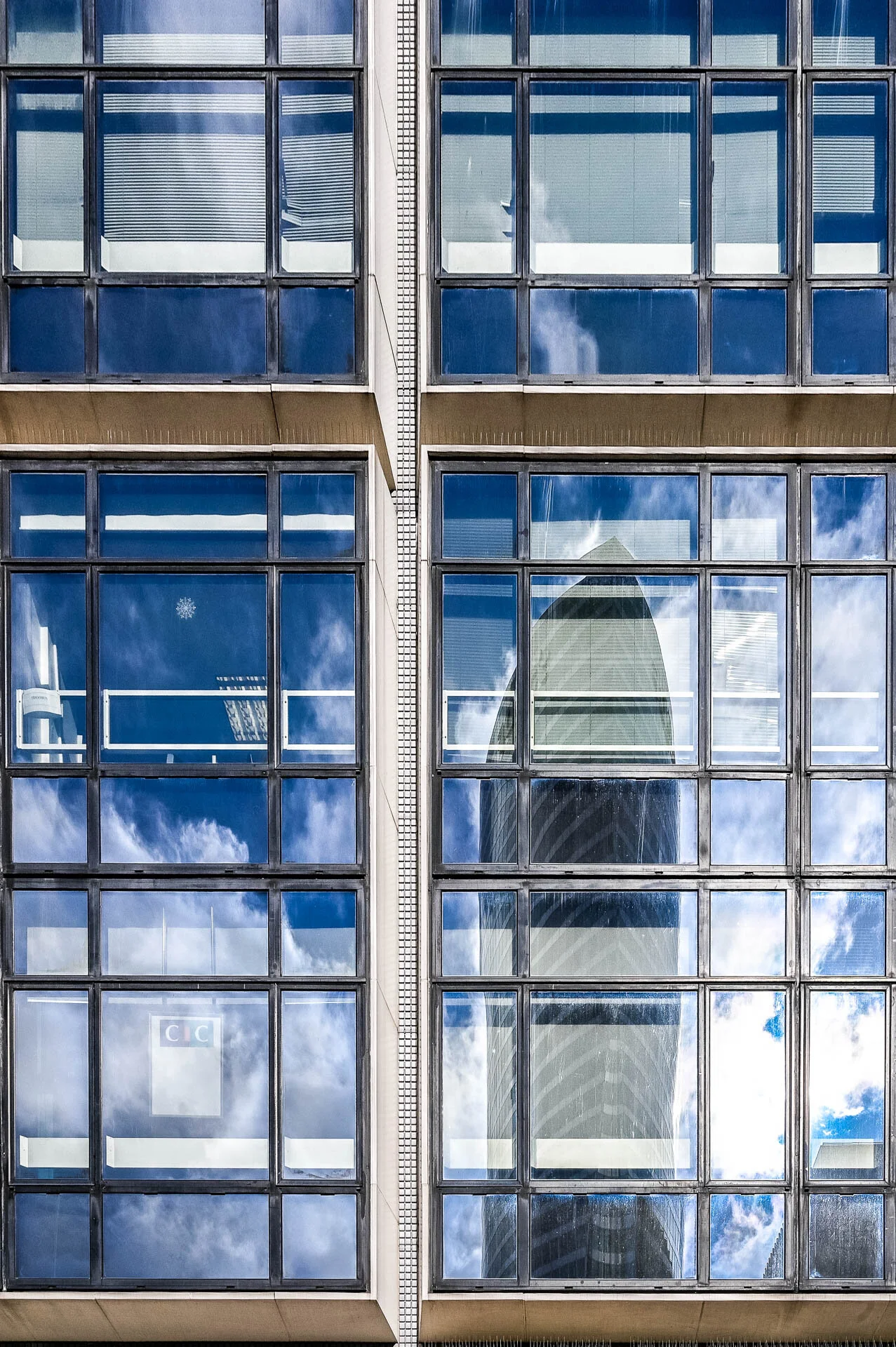 Reflets du ciel et d’un immeuble voisin dans les fenêtres d’une façade vitrée.
