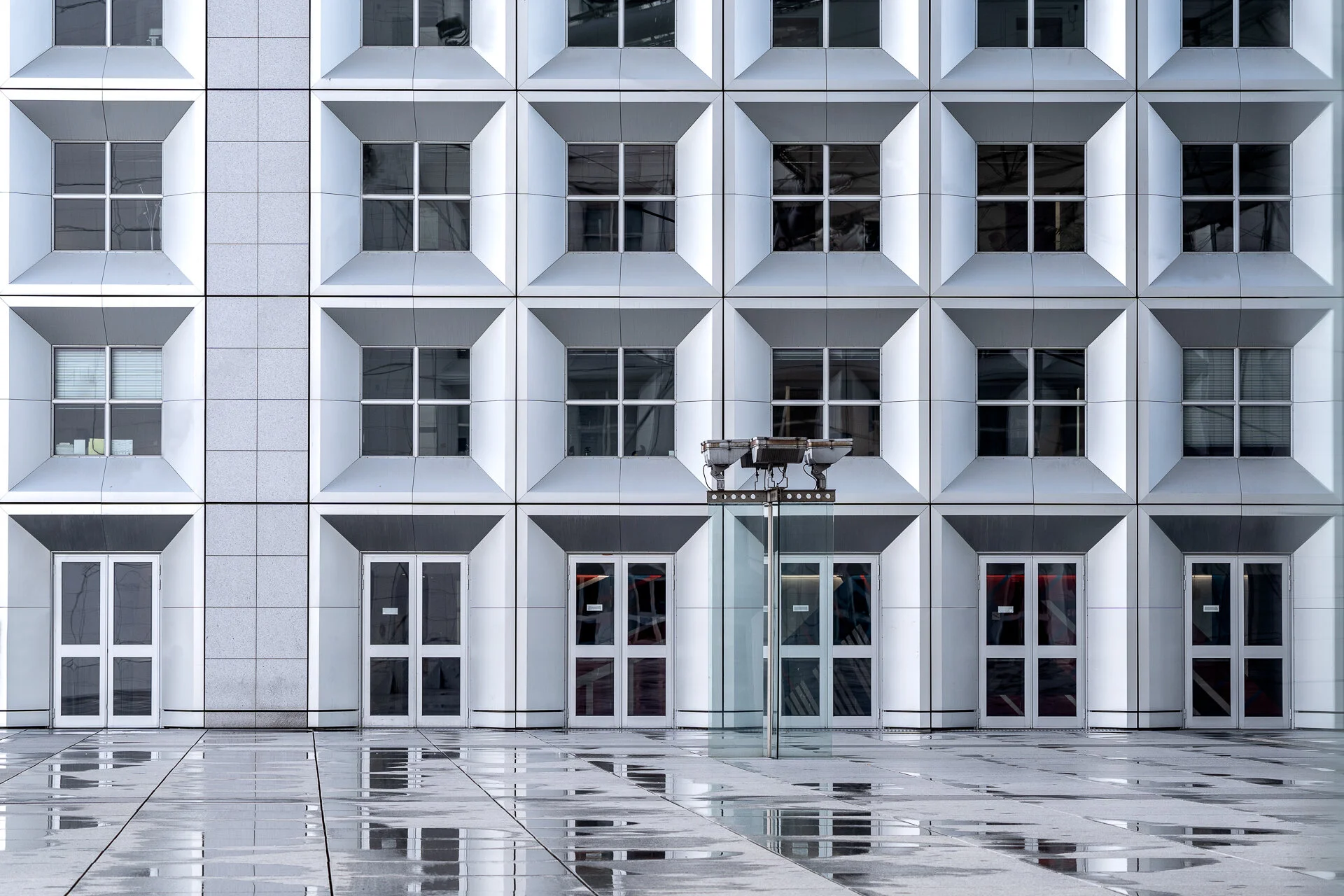 Façade géométrique d’un bâtiment moderne, avec des fenêtres encadrées de blanc et des reflets sur le parvis.