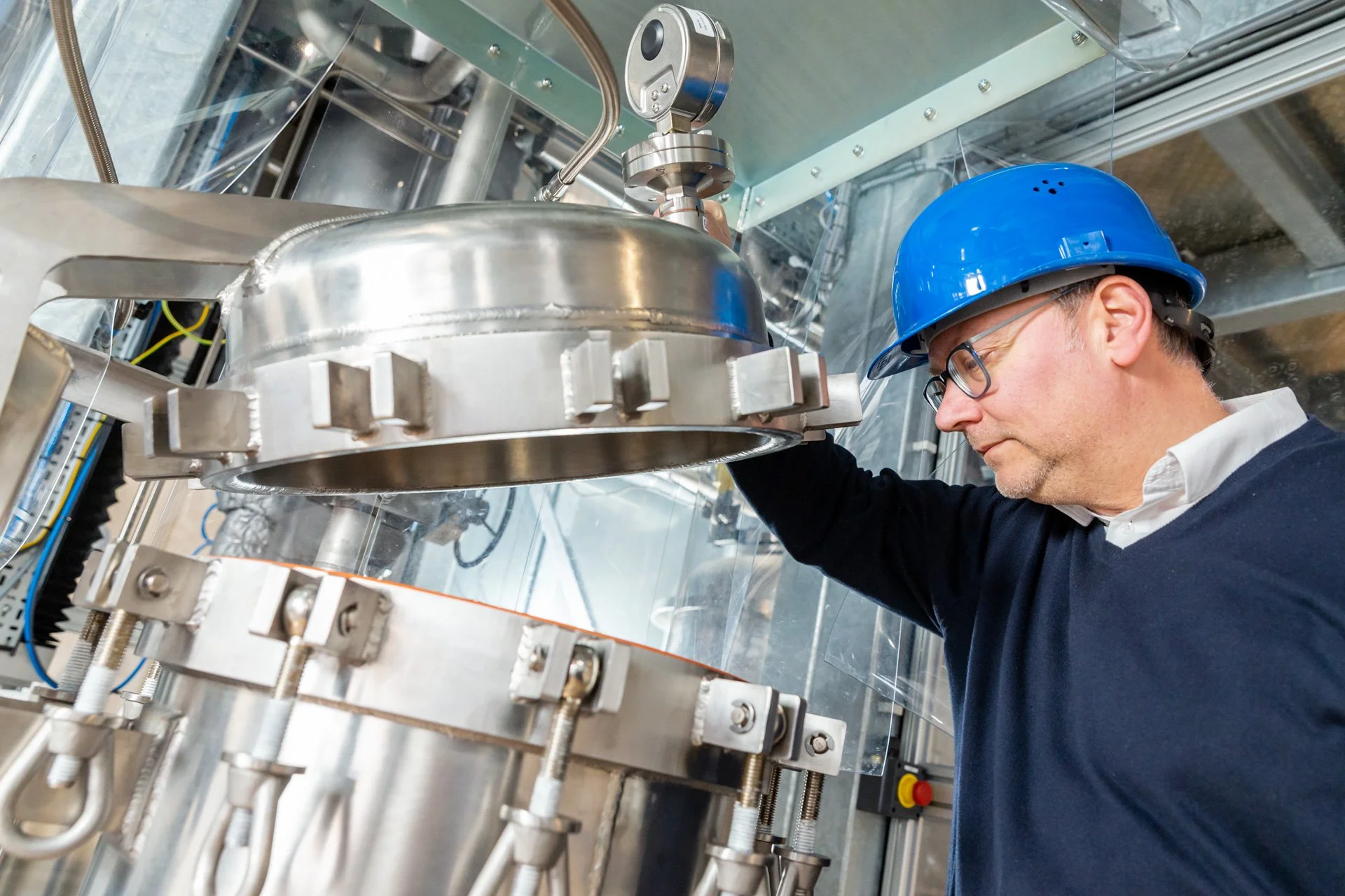 Un homme portant un casque de sécurité bleu et des lunettes examine une grande machine industrielle en métal.