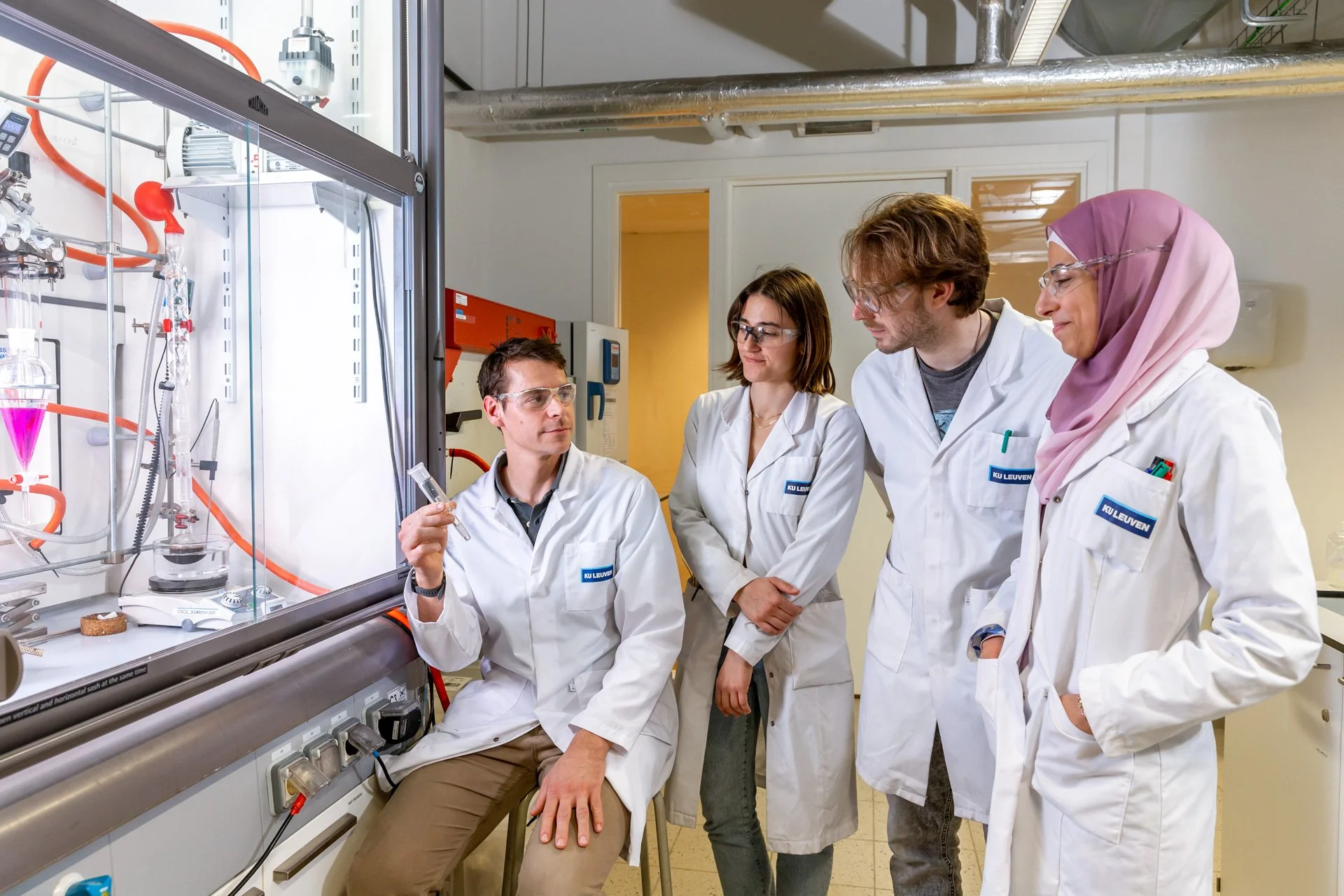 Quatre personnes en blouses blanches et lunettes de protection observent un équipement de laboratoire contenant des liquides colorés.
