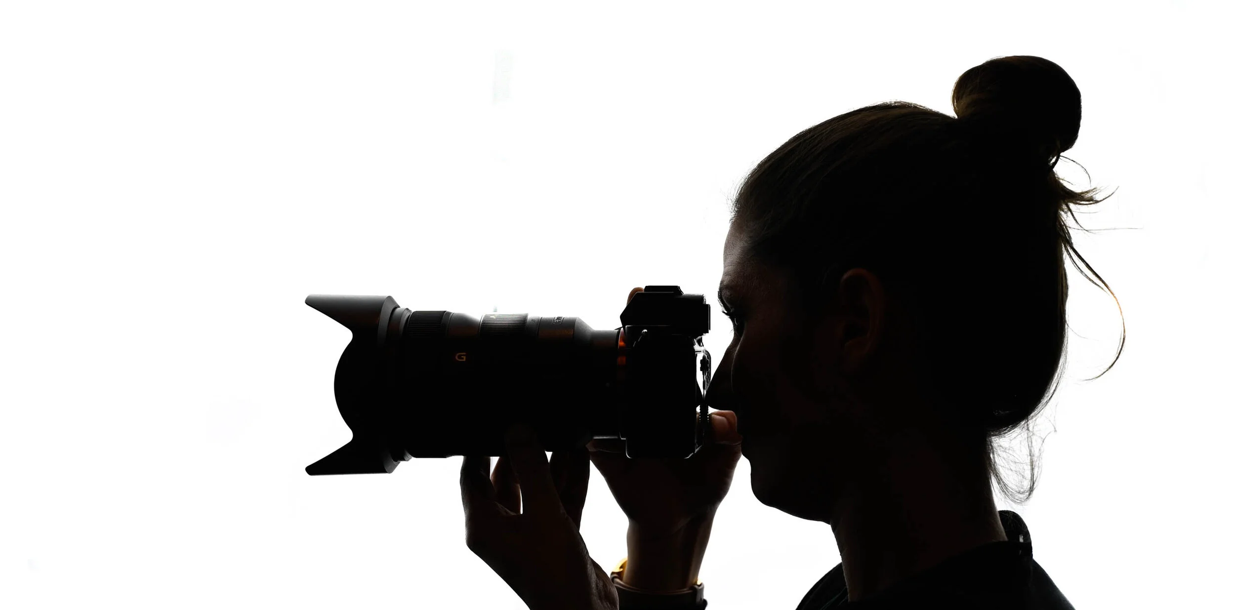 Silhouette de profil d’une photographe aux cheveux attachés regardant dans le viseur de son appareil sur fond blanc.