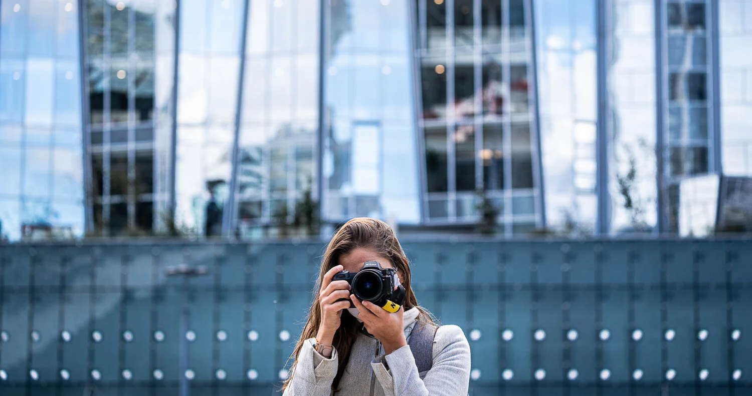 Une photographe tient son appareil devant son visage, avec la façade vitrée d’un bâtiment à l’arrière-plan.