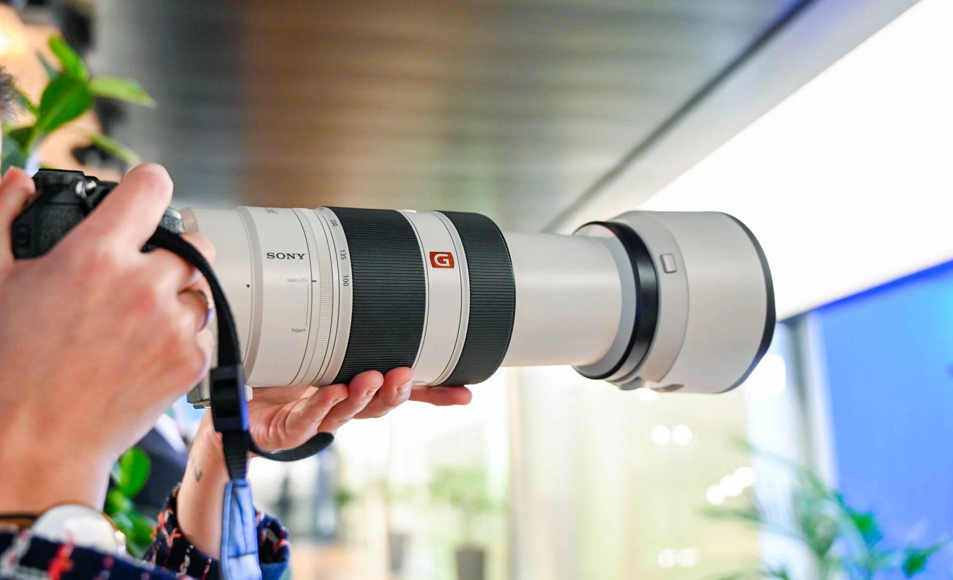 Gros plan sur les mains d’un photographe tenant un appareil équipé d’un long téléobjectif blanc.