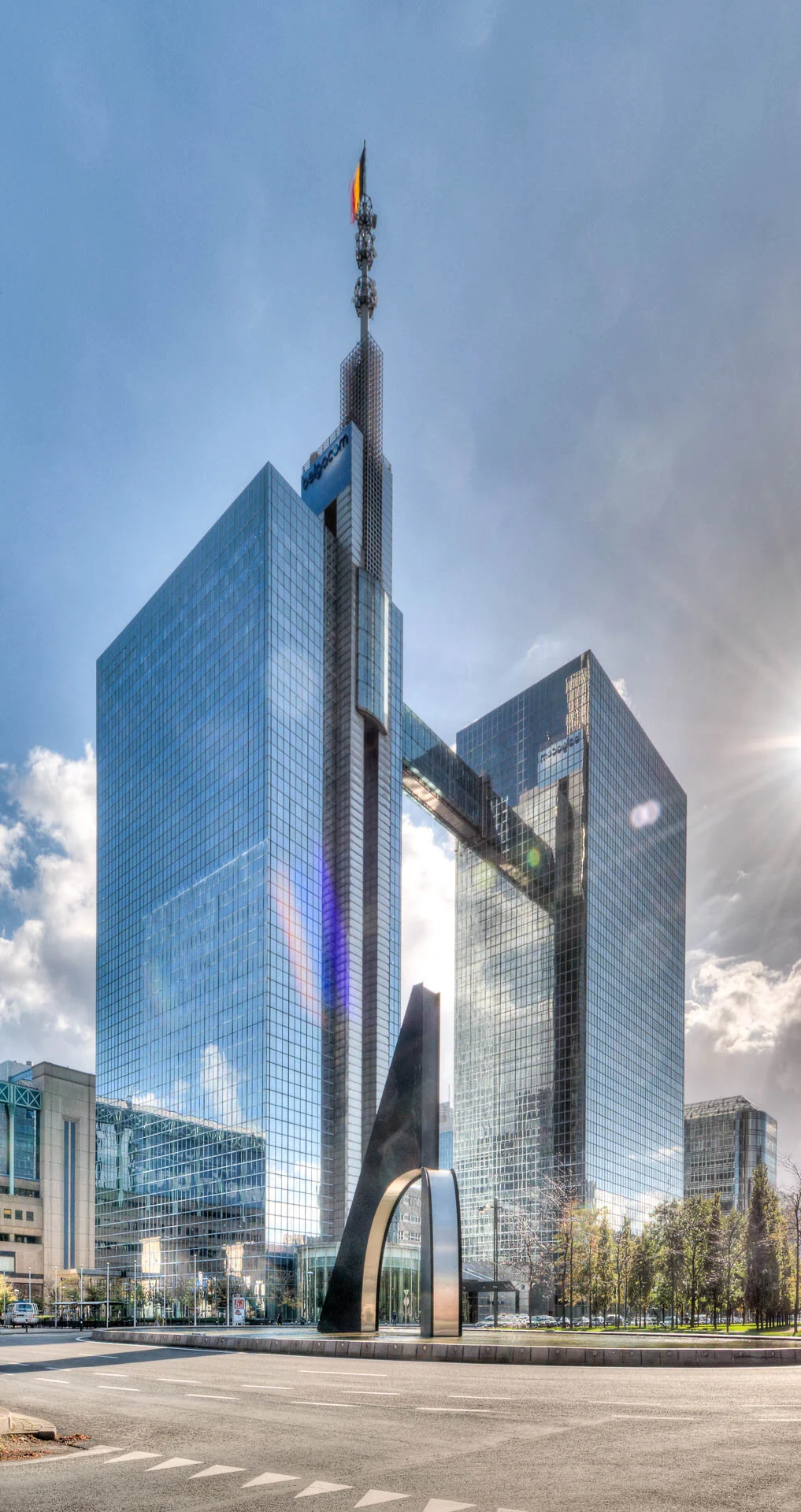 Vue d'ensemble d'un complexe de bureaux en verre avec une tour centrale surmontée d'une antenne et d'un drapeau, reliée à deux ailes par un pont aérien, et une sculpture métallique en forme d'arche au premier plan.
