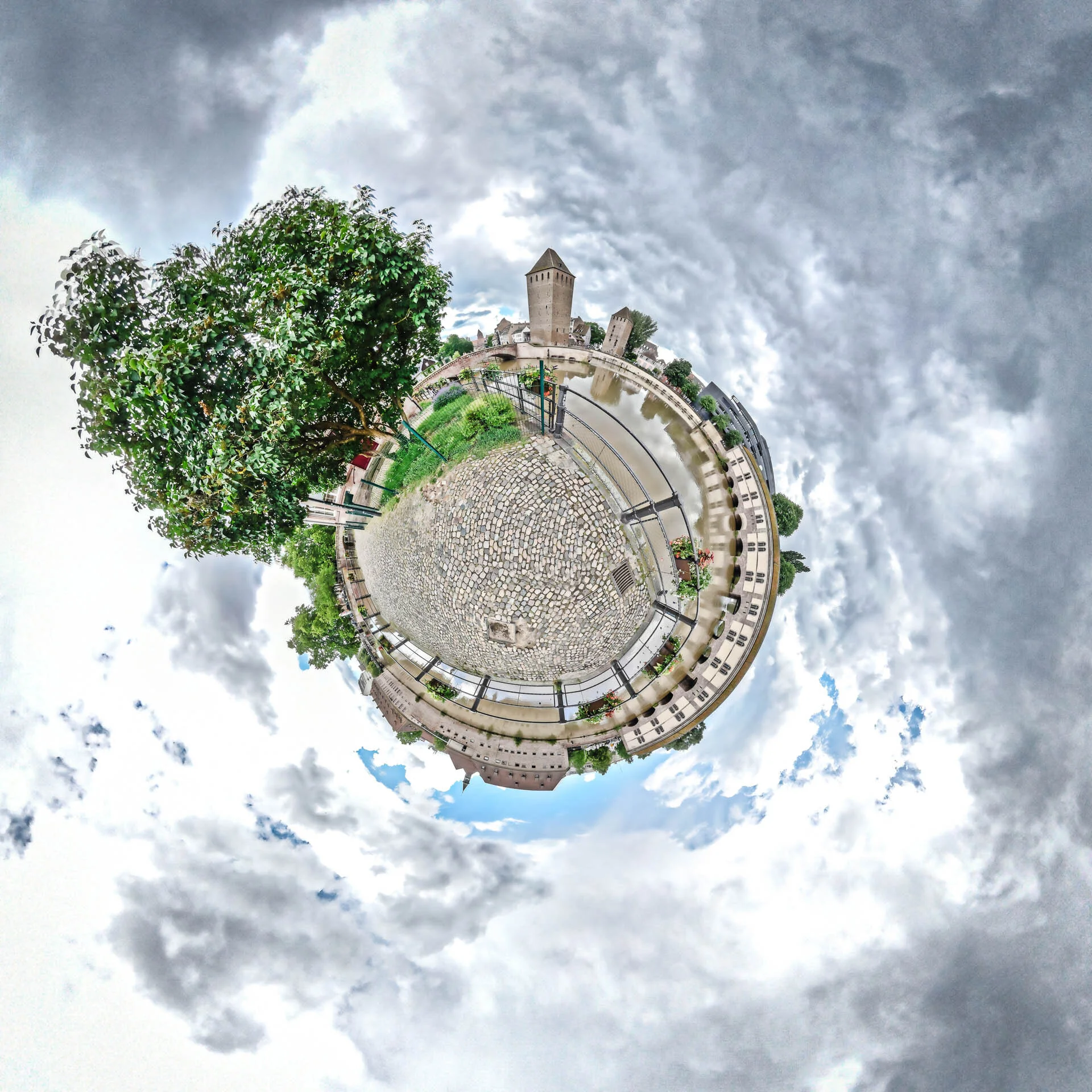 Mini-planète d’une place pavée à Strasbourg, entourée de bâtiments et d’arbres sous un ciel nuageux.