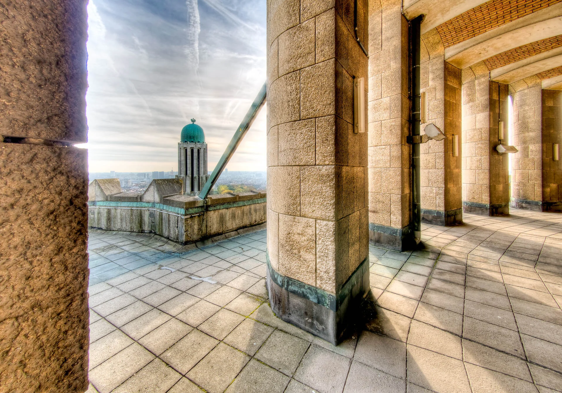 Vue d'un porche en pierre avec des colonnes massives et un sol carrelé, offrant une perspective sur une coupole verte et un paysage urbain au loin.