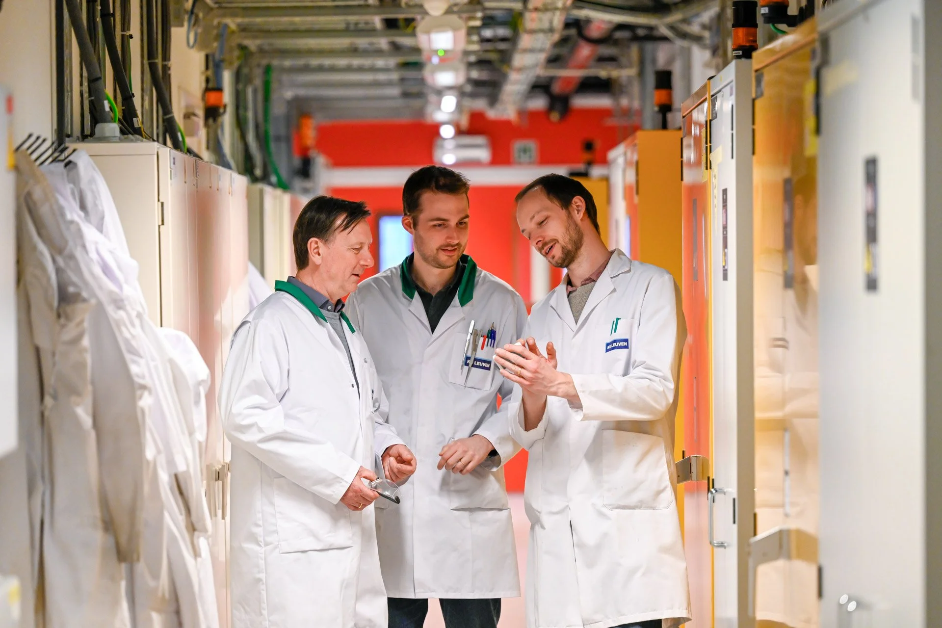 Trois personnes en blouses blanches discutent dans un couloir industriel ou de laboratoire.