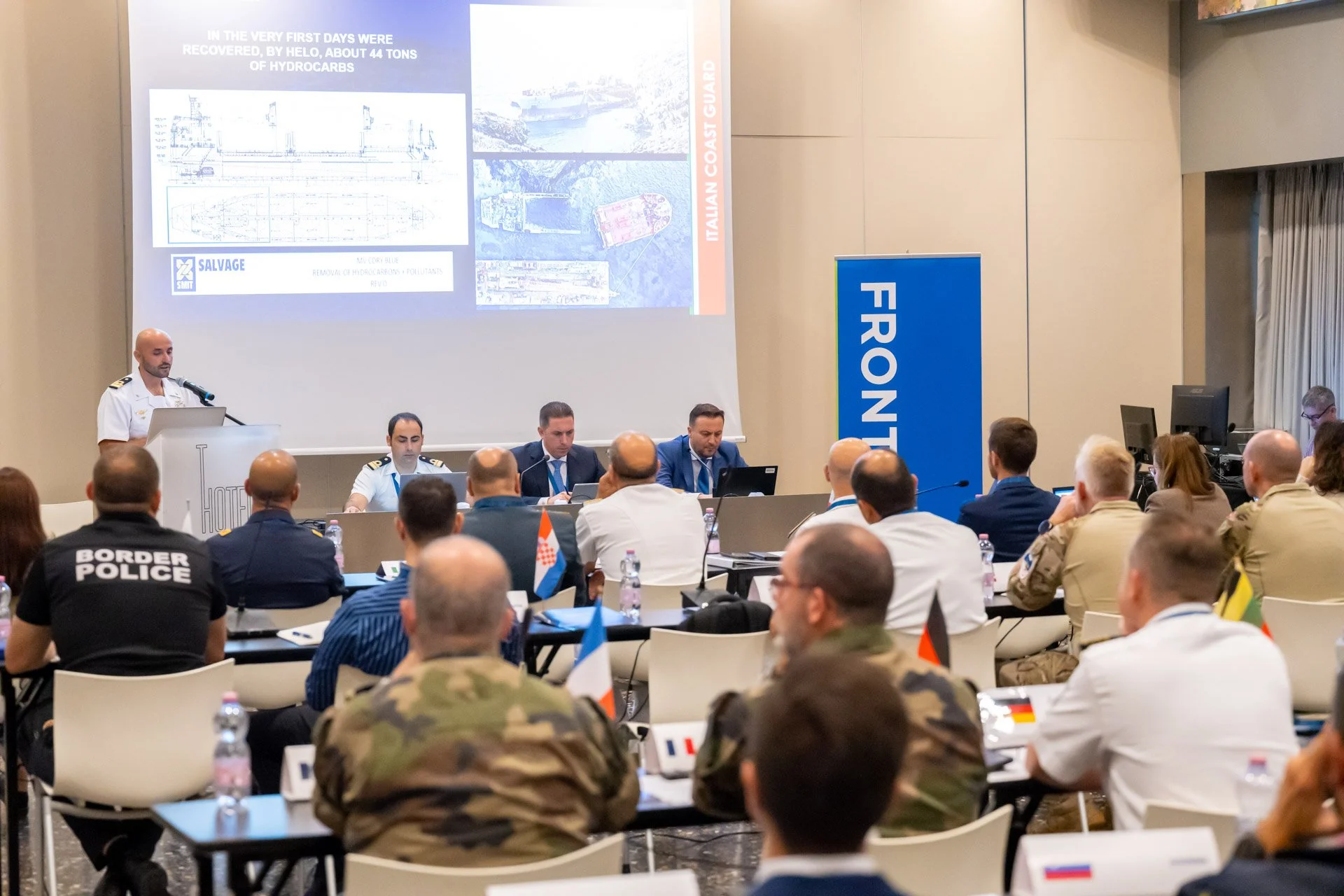 Un intervenant au pupitre s’adresse à des participants en tenue civile et en uniforme, devant un écran de projection et une bannière Frontex.