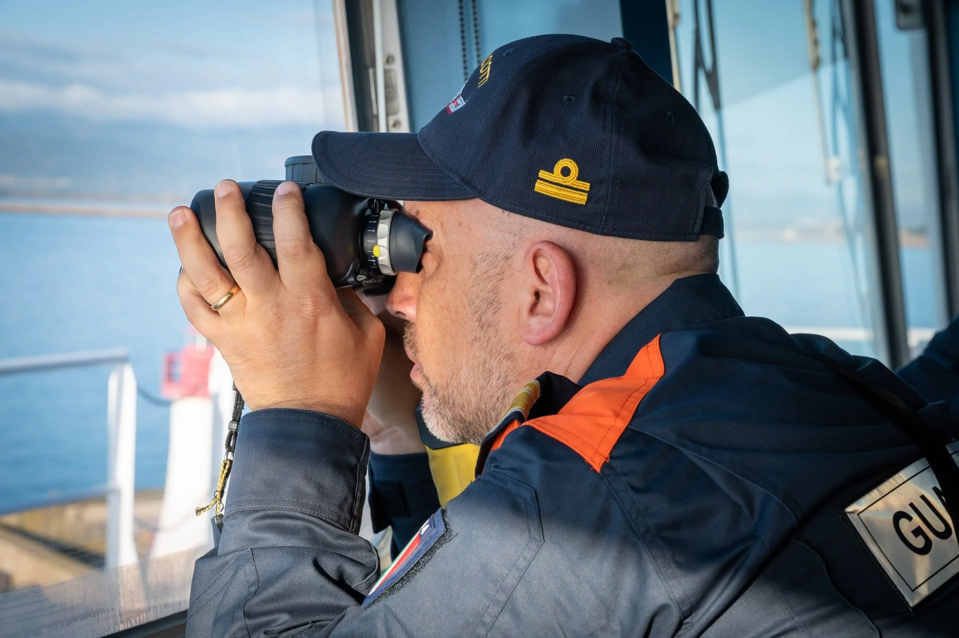 Un homme en uniforme observe la mer avec des jumelles depuis la passerelle d’un navire, lors d’un reportage pour Frontex.