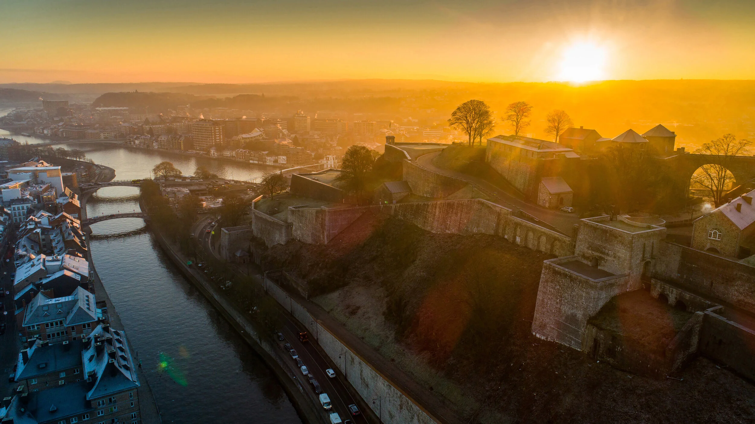 Vue aérienne d’une ville traversée par une rivière, avec une citadelle sur les hauteurs au coucher du soleil.