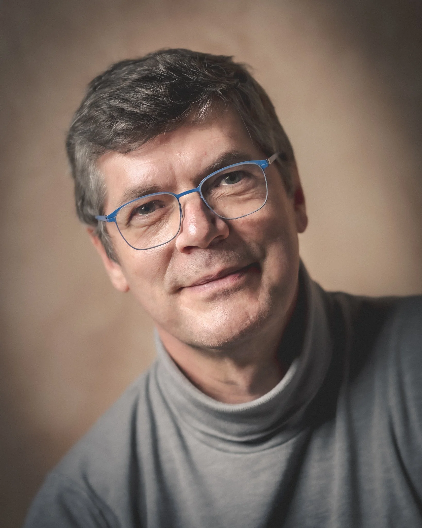 Portrait de Paul Marnef portant des lunettes bleues et un pull gris, devant un fond brun.