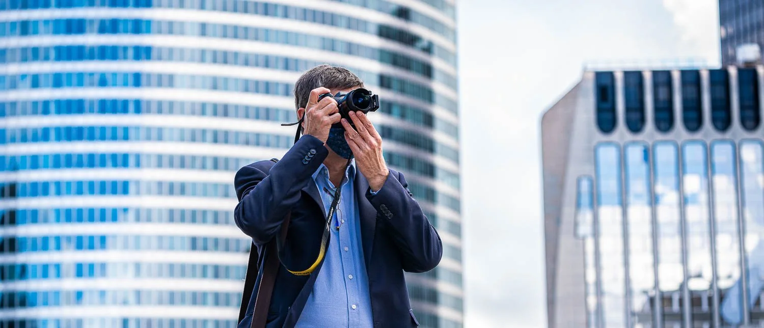 Un photographe tient son appareil devant son visage, avec des immeubles modernes à l’arrière-plan.
