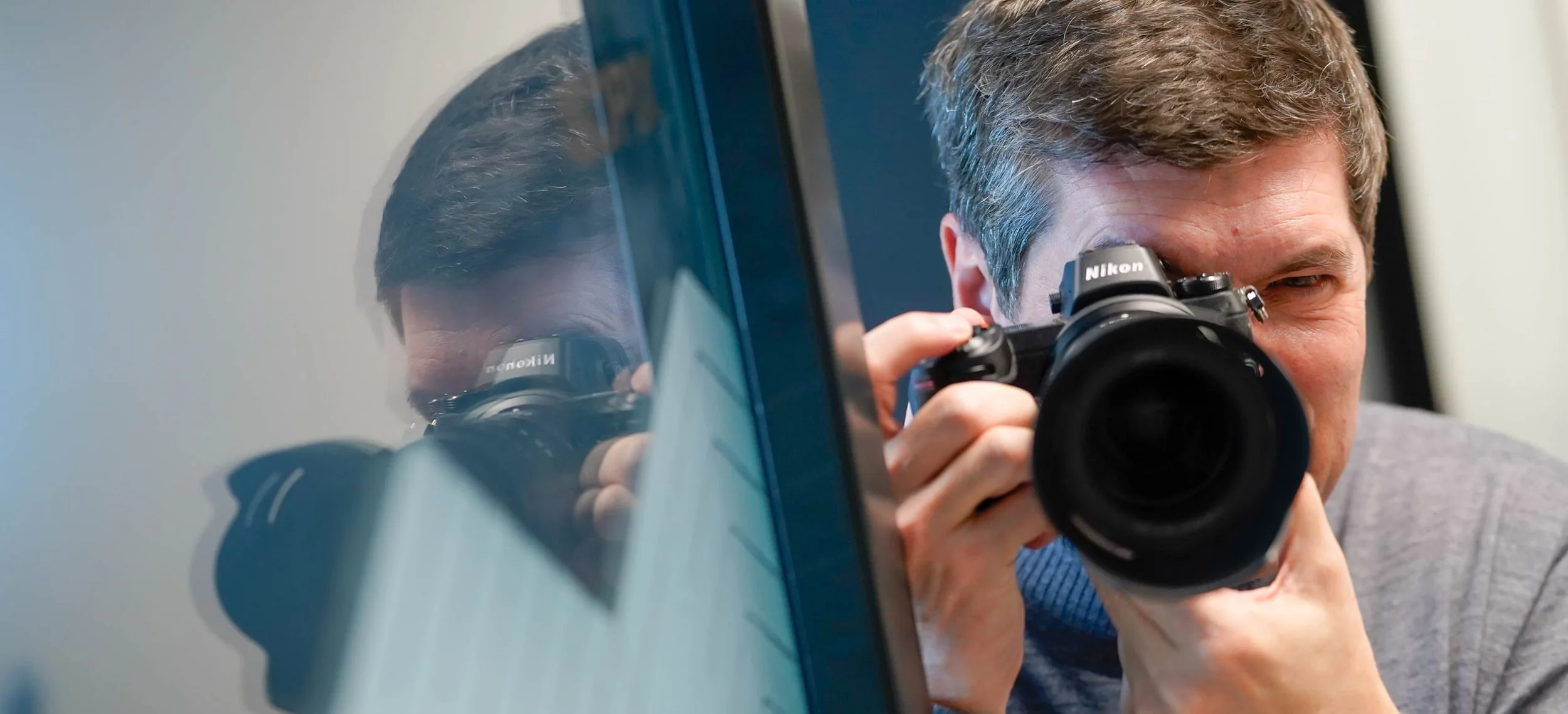 Un photographe vise avec son appareil devant une vitre reflétant son visage et son équipement.