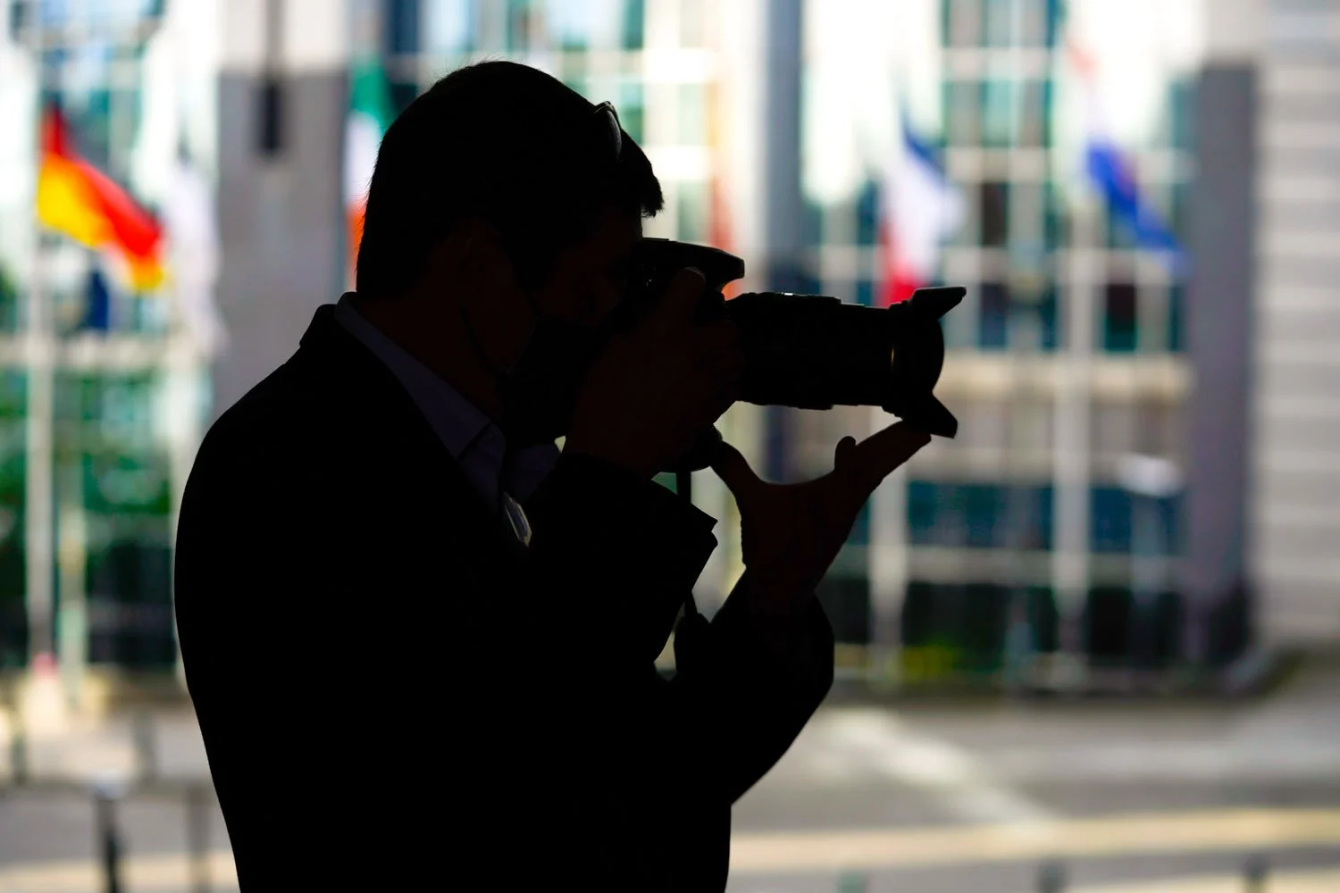 Photographe en silhouette prenant une photo, avec des drapeaux et des immeubles flous en arrière-plan.