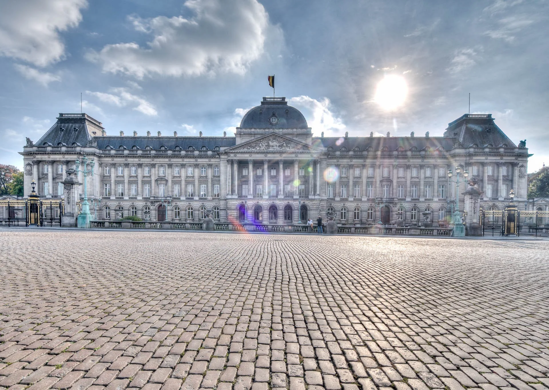 Le Palais royal de Bruxelles photographié au ras des pavés, sous un ciel nuageux traversé de lumière.