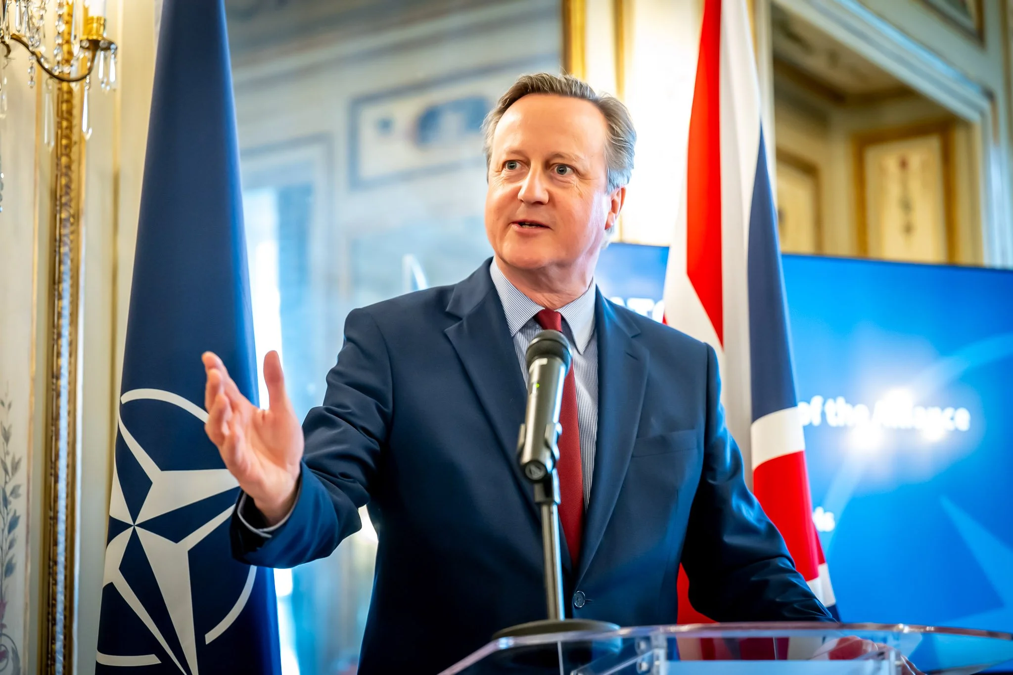 David Cameron devant un micro avec drapeaux de l'OTAN et du Royaume-Uni en arrière-plan.