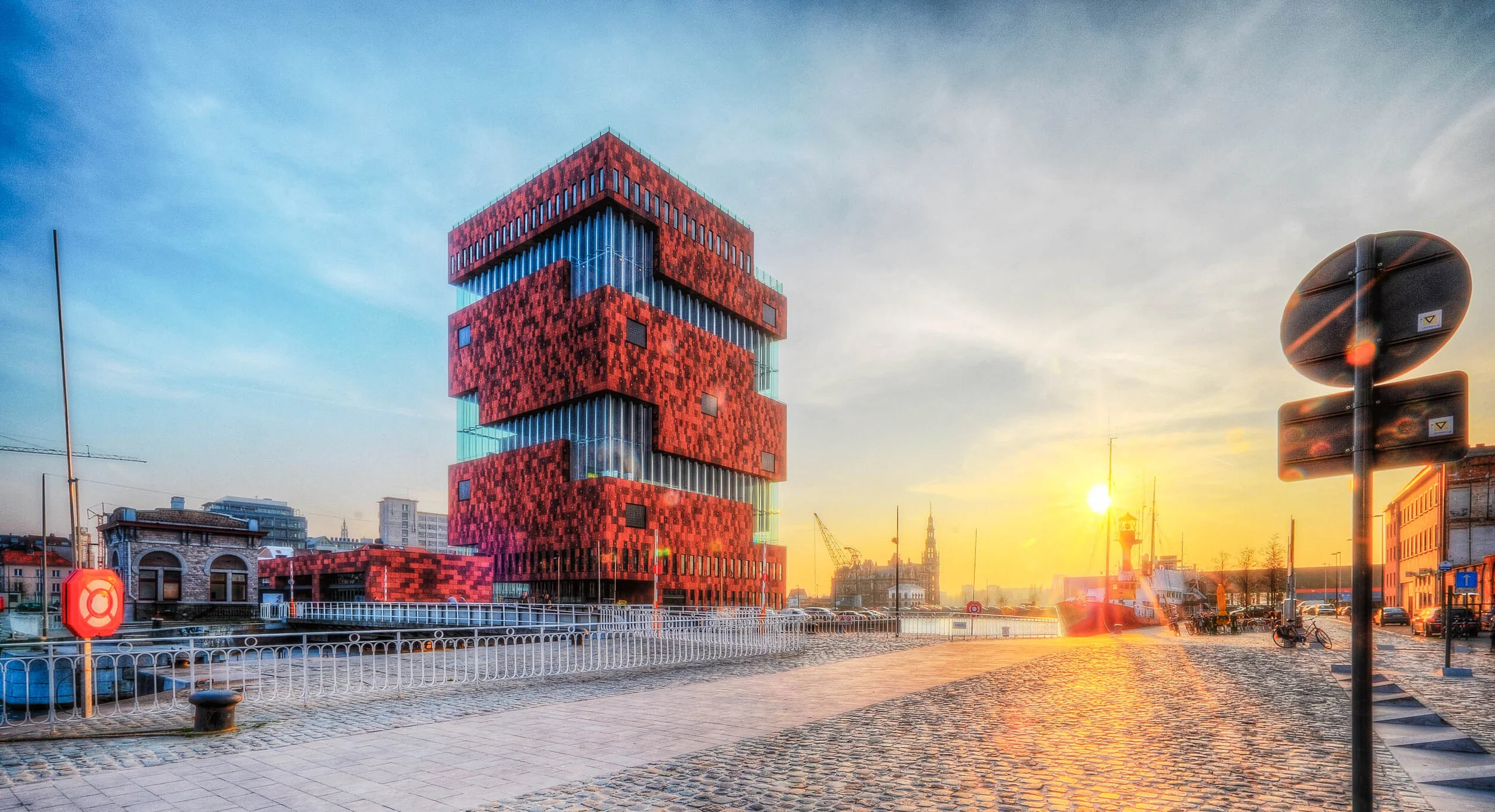 Bâtiment aux volumes rouges empilés, au bord de l’eau au coucher du soleil.