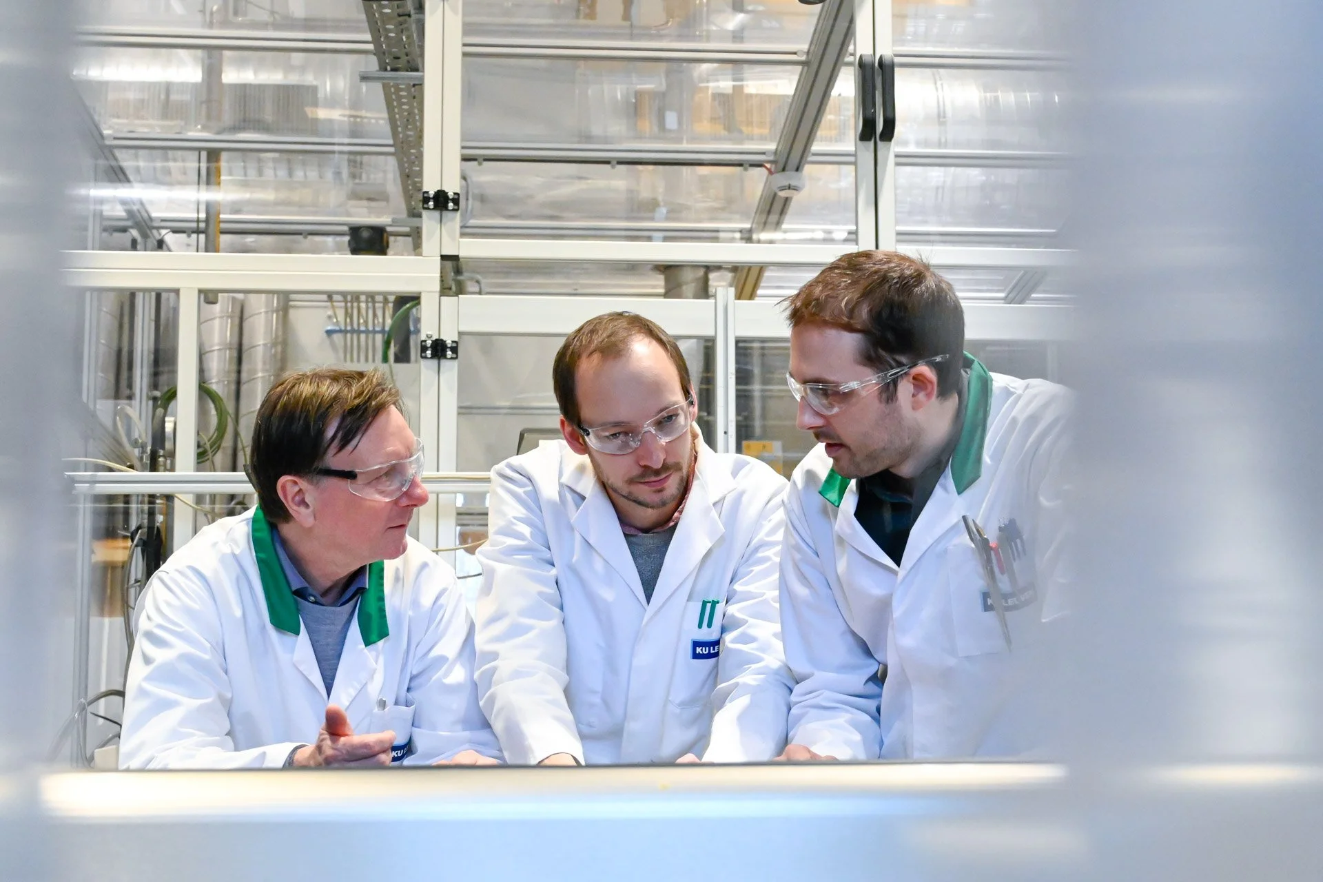 Trois personnes portant des blouses blanches et des lunettes de protection discutent ensemble dans un environnement industriel ou de laboratoire.