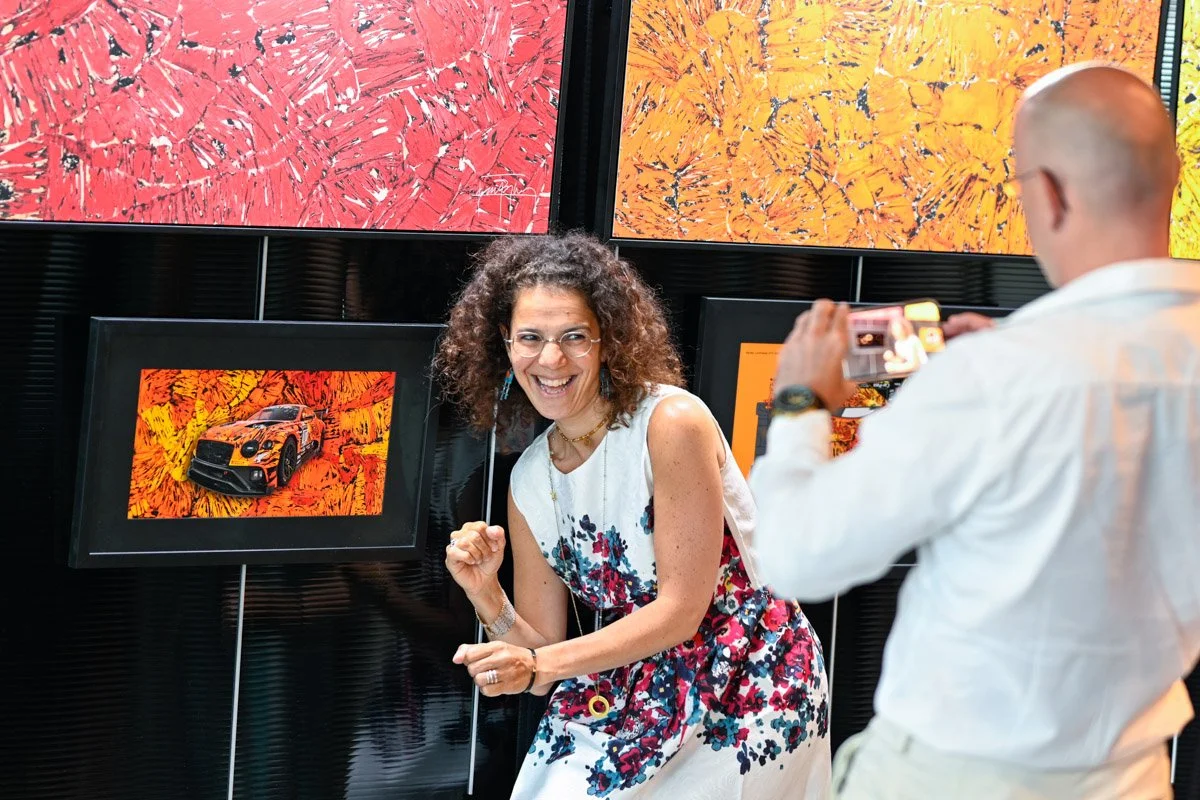 Un participant photographie avec son téléphone une femme souriante devant des tableaux colorés lors d’un événement.