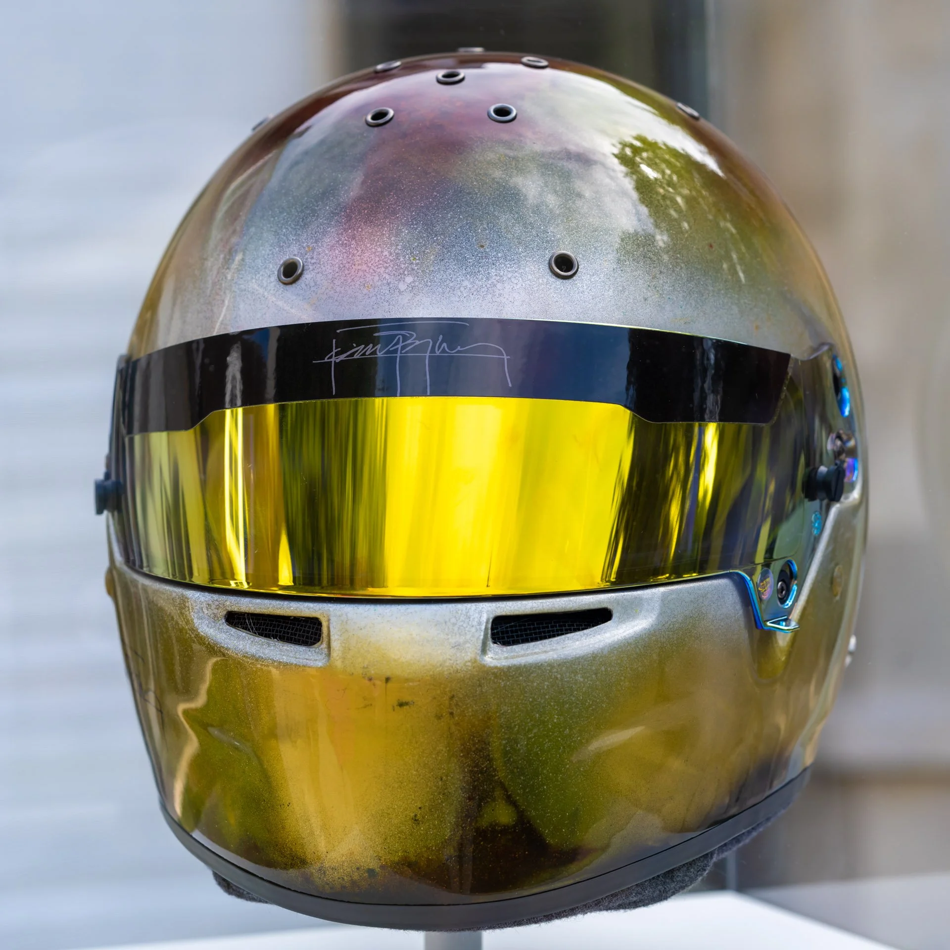Casque de pilote de course avec visière dorée, reflet d'arbres sur la surface, signatures et boutons latéraux.