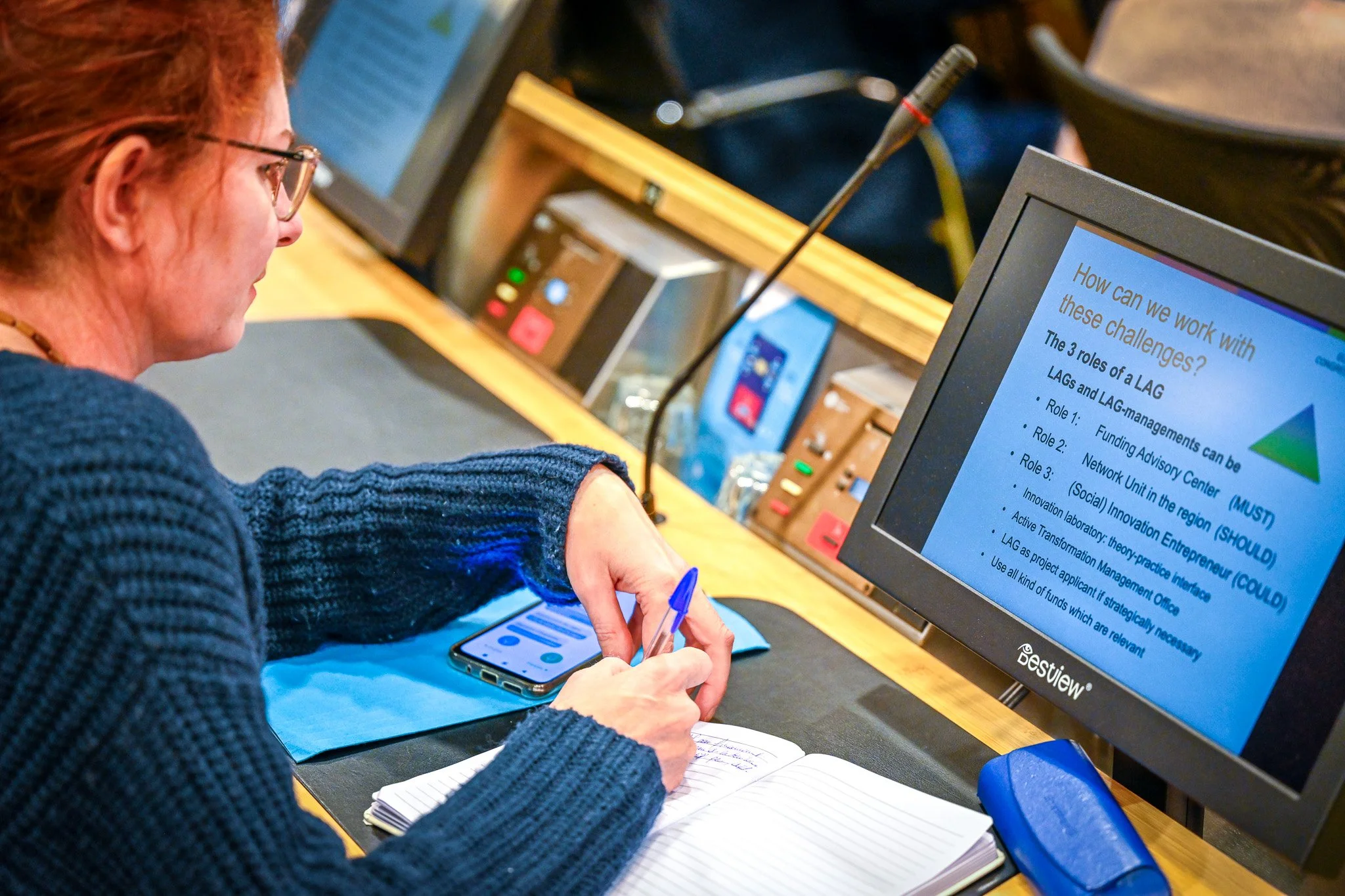 Une femme assise à une table, écrivant dans un carnet, avec une tablette et un ordinateur portable devant elle. L'écran de l'ordinateur affiche un document avec des points de liste sur la gestion des défis liés aux connaissances. Il y a également un