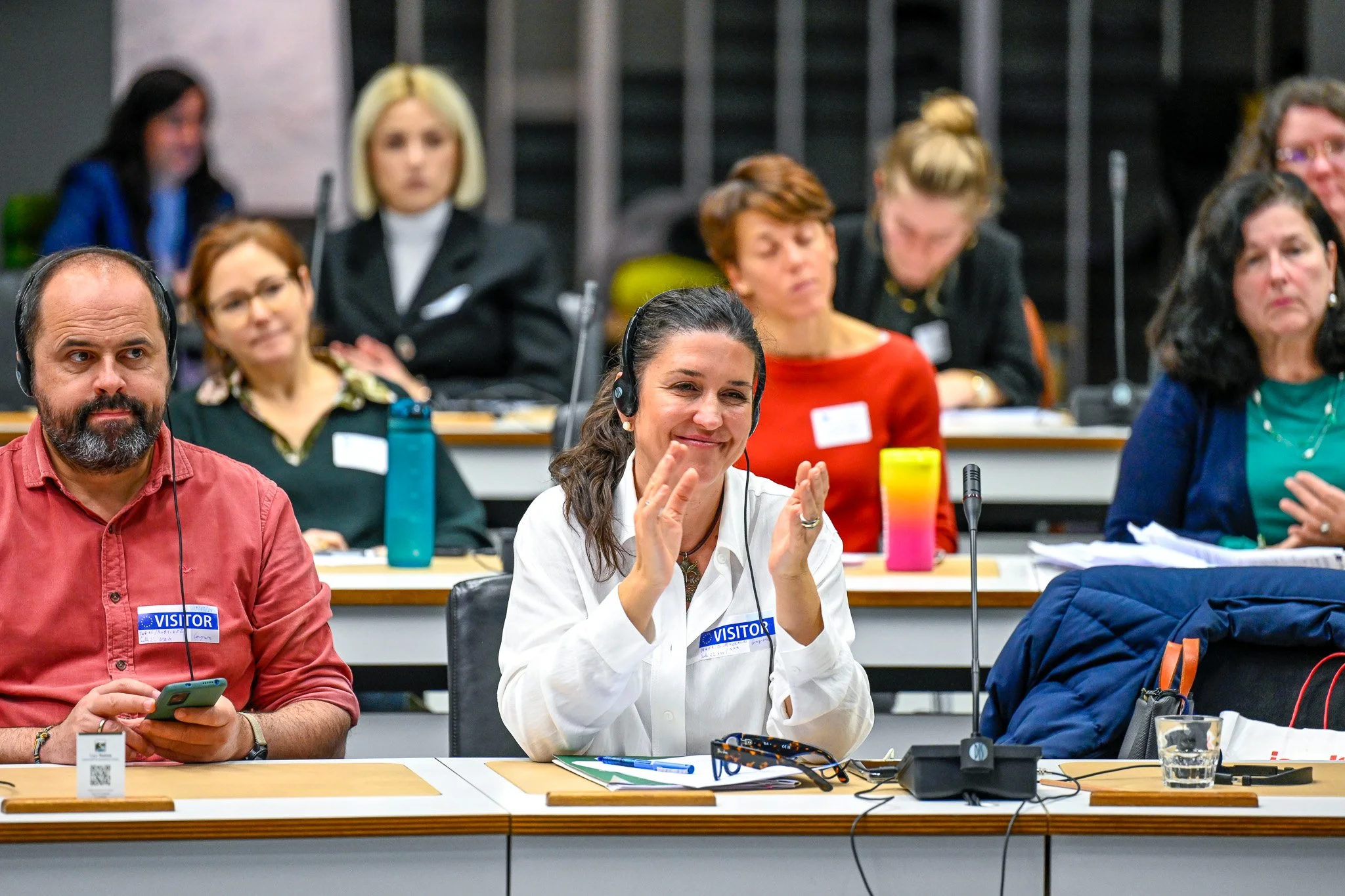 une conférence à Bruxelles. Groupe de personnes assises à des tables en conférence, certaines applaudissant, avec des écouteurs et des badges de visiteur.