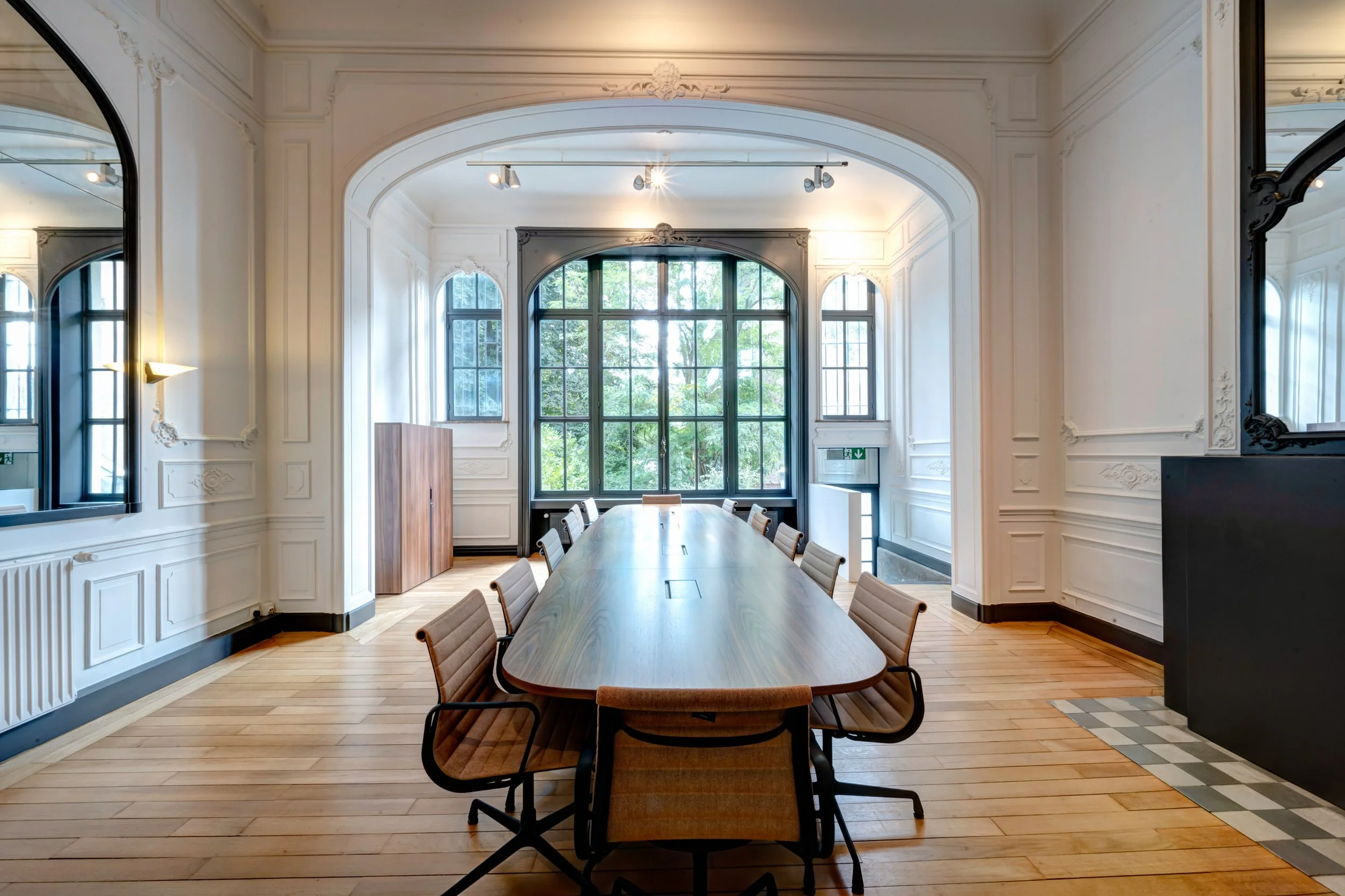 Salle de réunion avec une longue table en bois, des chaises et des murs blancs moulurés, éclairée par de grandes fenêtres.