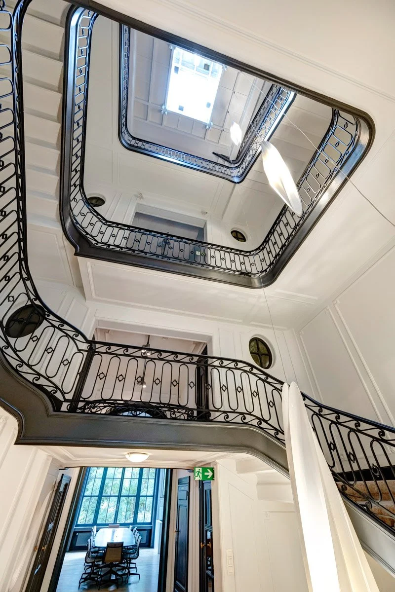 Vue en contre-plongée d'une cage d'escalier intérieure avec des balustrades en fer forgé noir, des murs blancs et un puits de lumière au plafond.