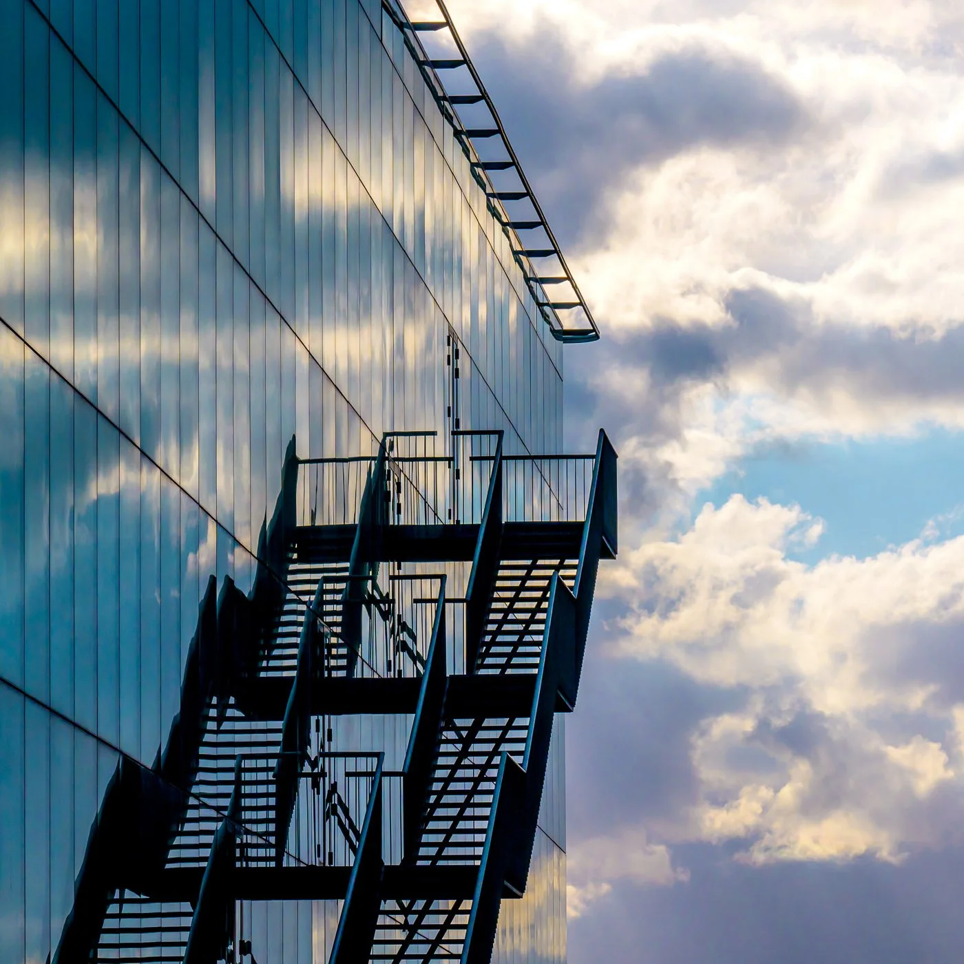 Façade vitrée d’un bâtiment à Louvain-la-Neuve, avec un escalier métallique extérieur et des nuages éclairés par le soleil.