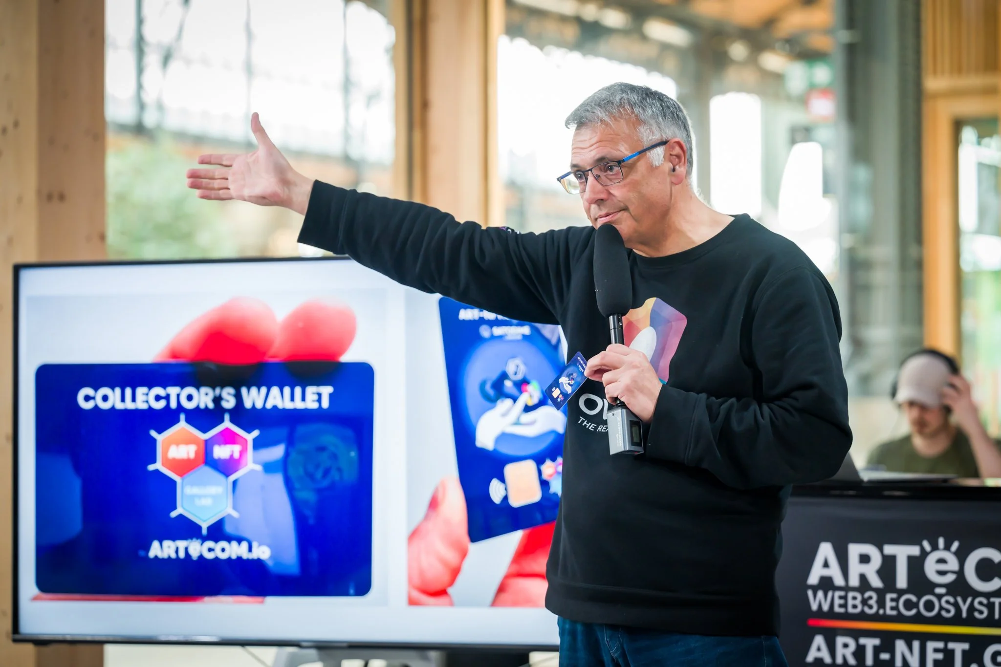 Un homme tient un micro et une carte bleue devant un écran affichant le logo Collector's Wallet et Art.com.io, lors d'une présentation sur un podium ArtéC.