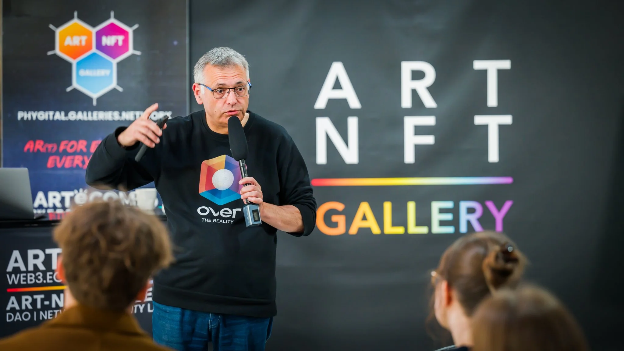 Un homme s’adresse au public avec un micro devant une bannière « ART NFT GALLERY ».
