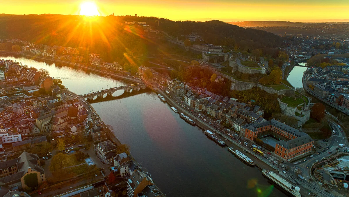 Vue aérienne d’une ville traversée par une rivière, avec un pont et une colline au coucher du soleil.