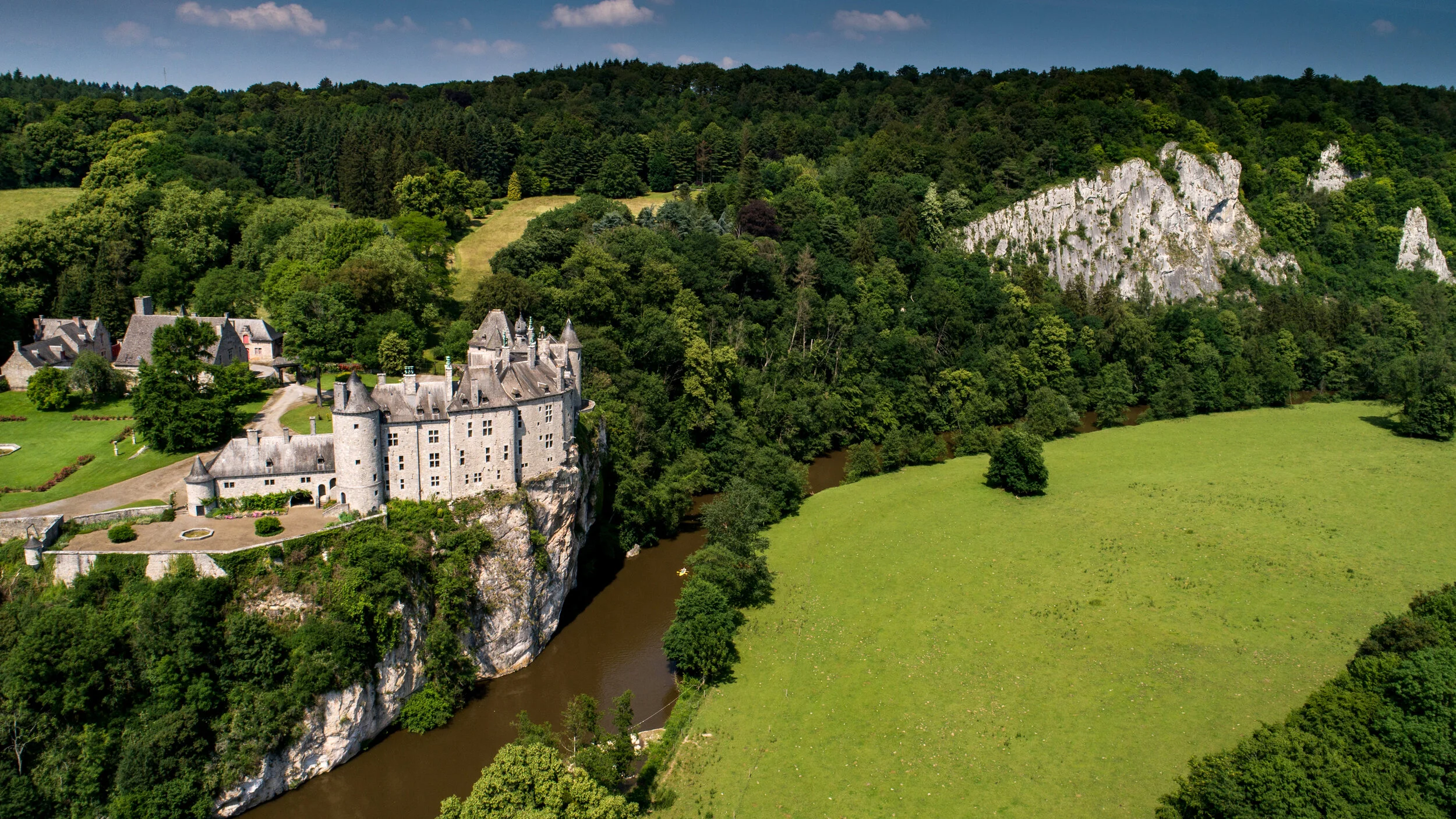 Vue aérienne d’un château au bord d’une rivière, au pied de falaises boisées.