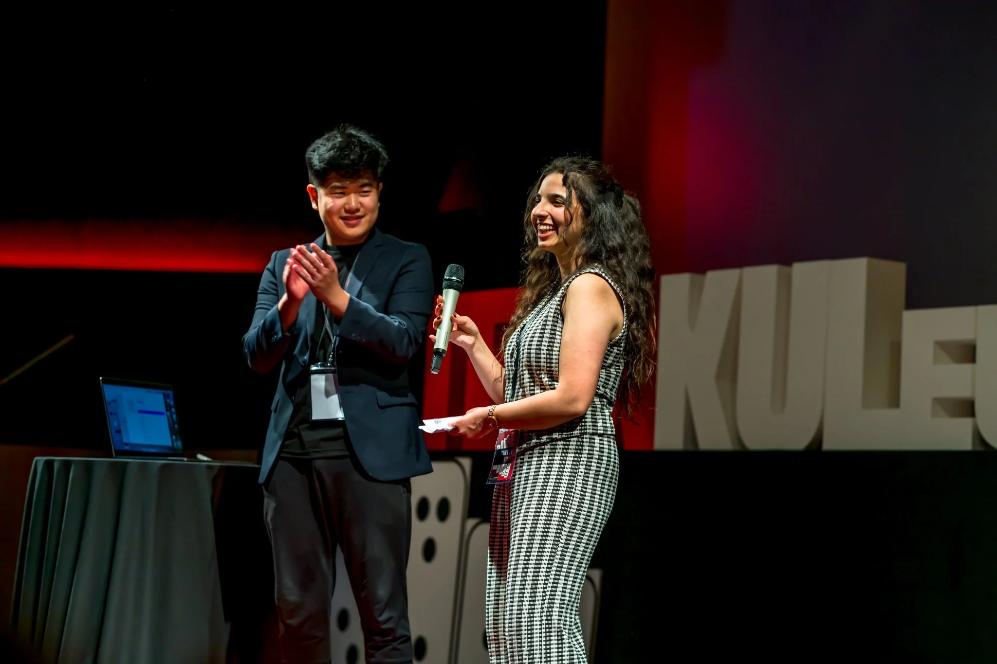 Un homme en costume applaudit à côté d'une femme en robe à carreaux qui tient un microphone sur une scène éclairée de rouge, avec un ordinateur portable posé sur une table à gauche.