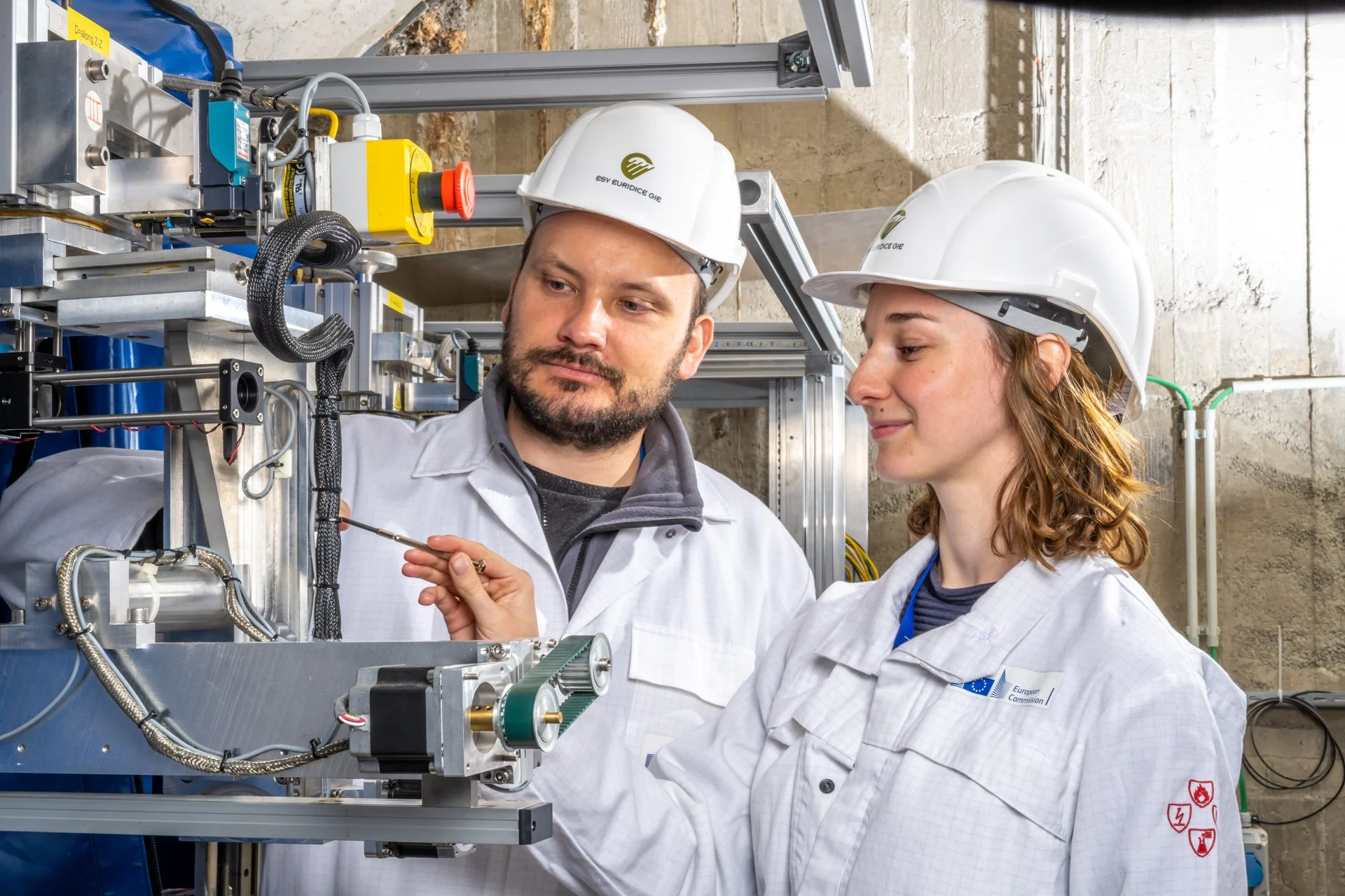 Deux techniciens en blouses blanches et casques de sécurité examinent une machine industrielle.