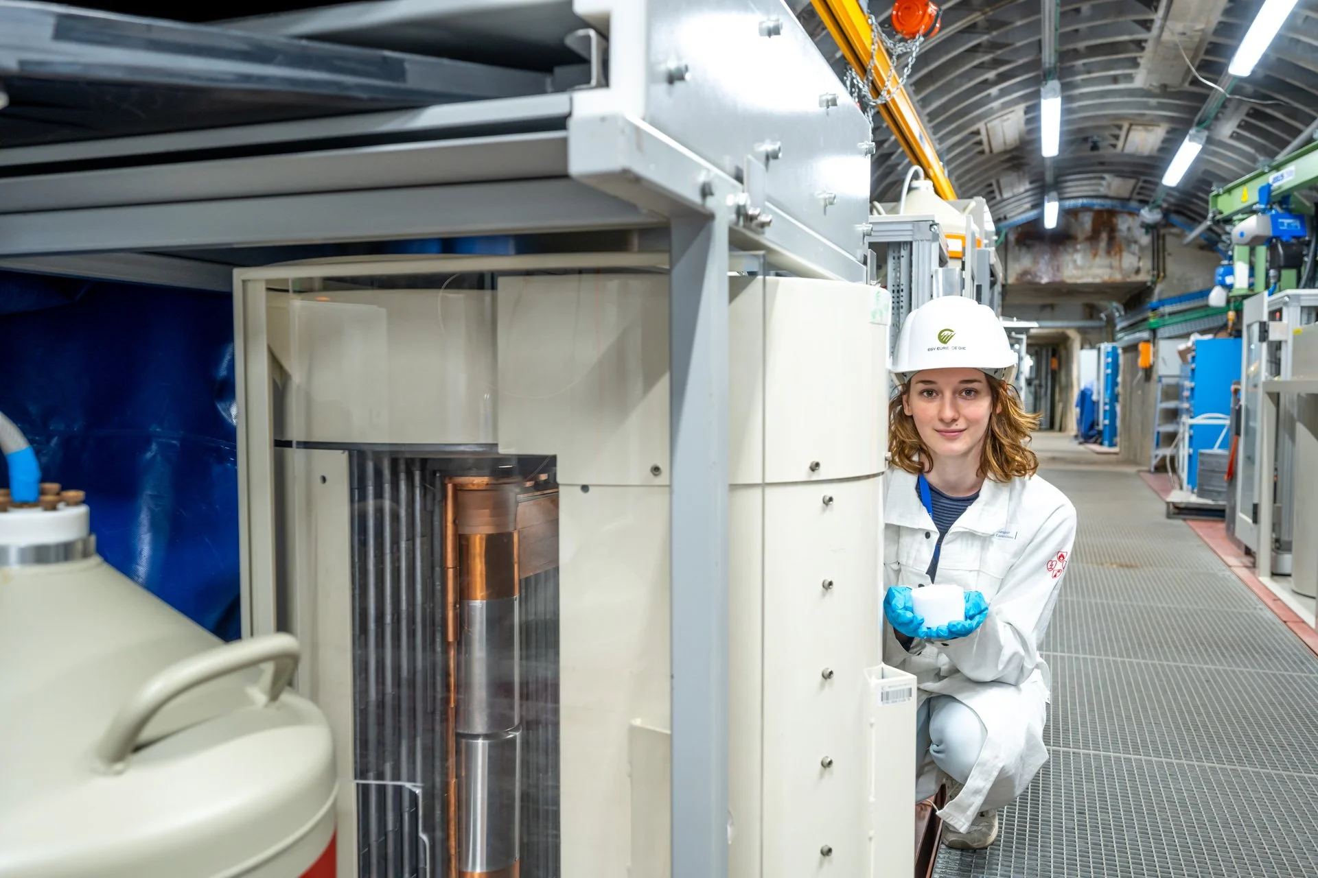 Femme en combinaison de protection blanche et casque de chantier accroupie à côté d'un équipement industriel complexe.