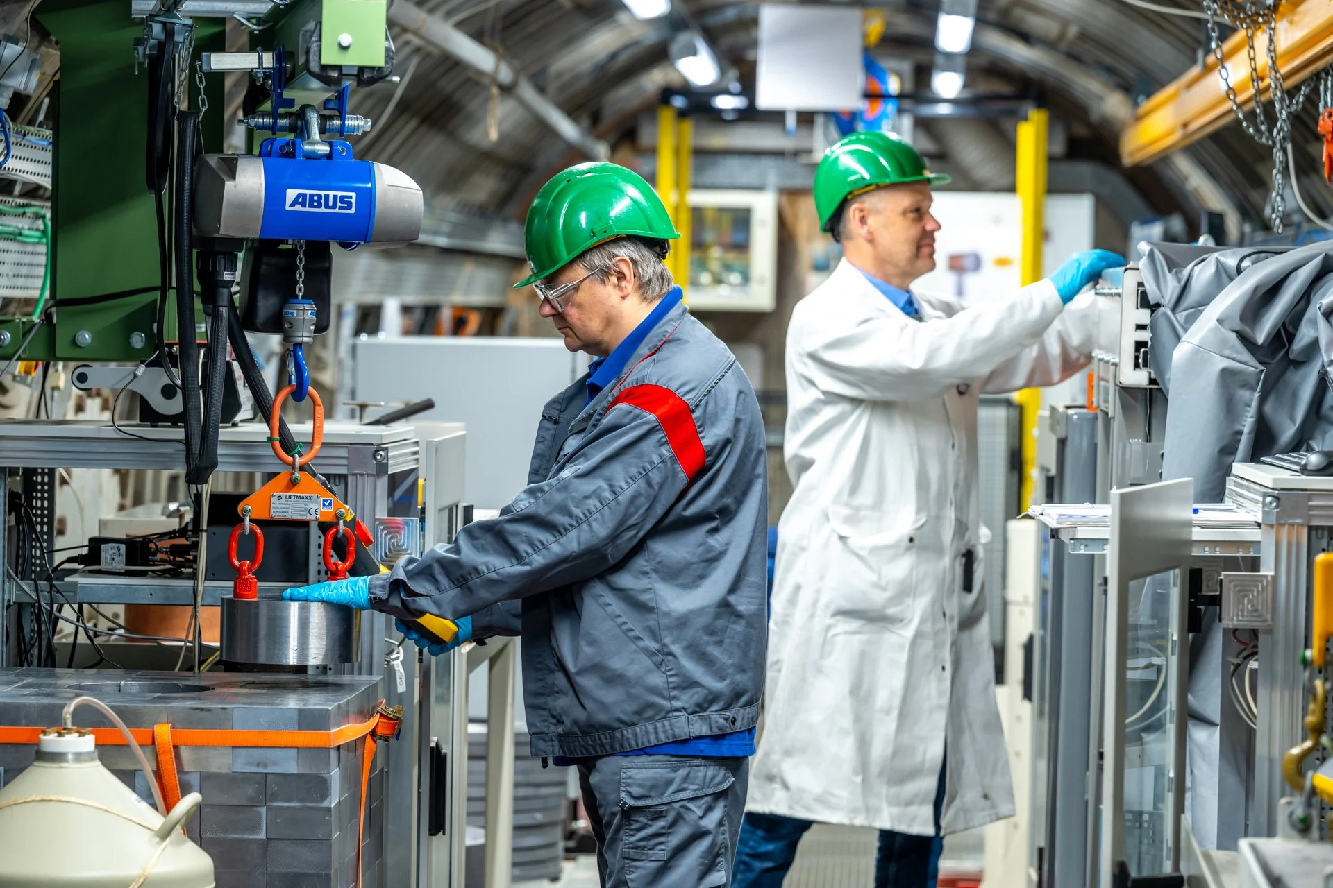Deux personnes portant des casques verts travaillent parmi des équipements scientifiques et des conduites dans une installation du JRC à Geel.