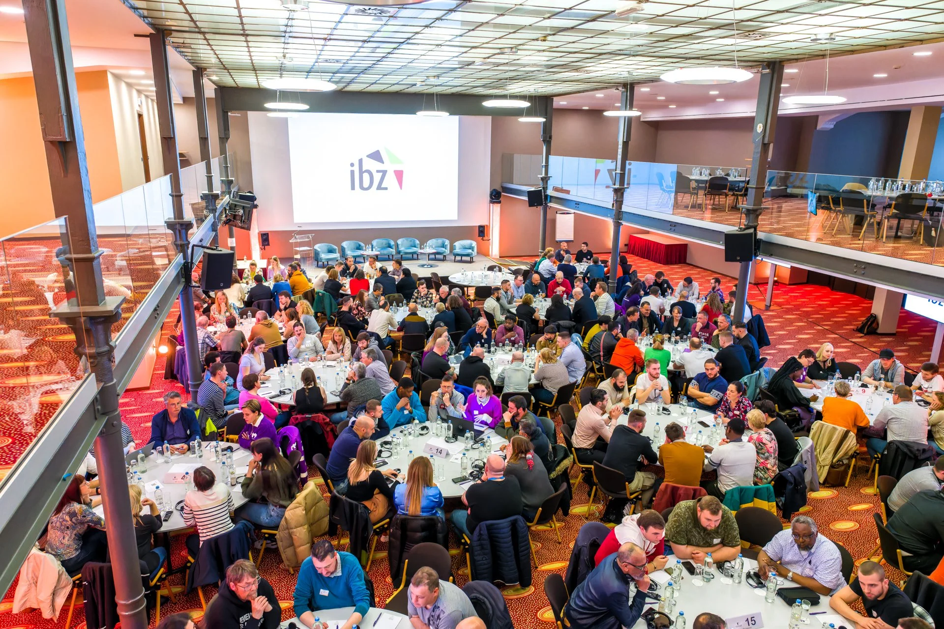 Participants assis autour de tables rondes dans une grande salle, devant un écran affichant le logo « ibz ».