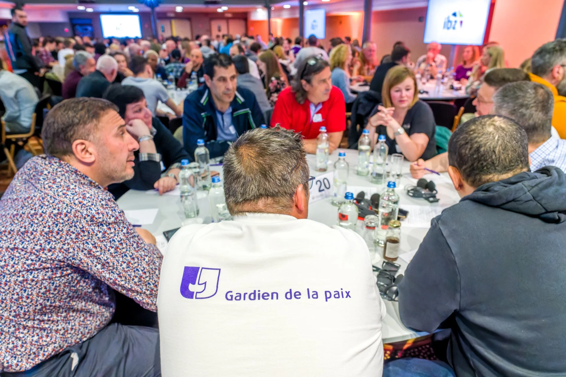 Participants échangeant autour d’une table avec des bouteilles d’eau et des microphones, dont un homme portant un t-shirt « Gardien de la paix ».