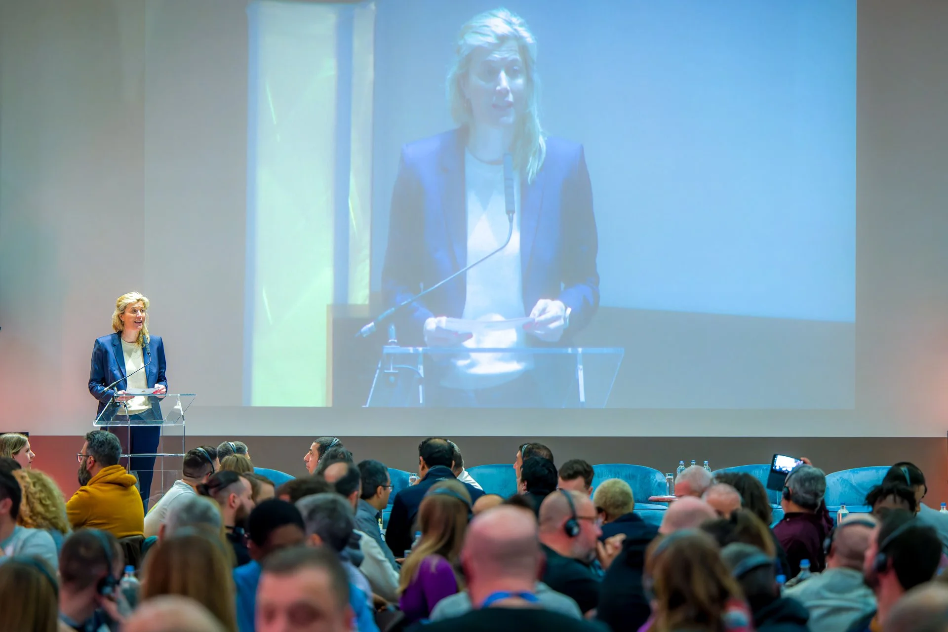 une conférence où l'on voit une personnalité prendre la parole. Une femme en costume parle à une conférence, debout à un pupitre avec un microphone. Elle est projetée sur un grand écran derrière elle. Un public de personnes assises écoute att