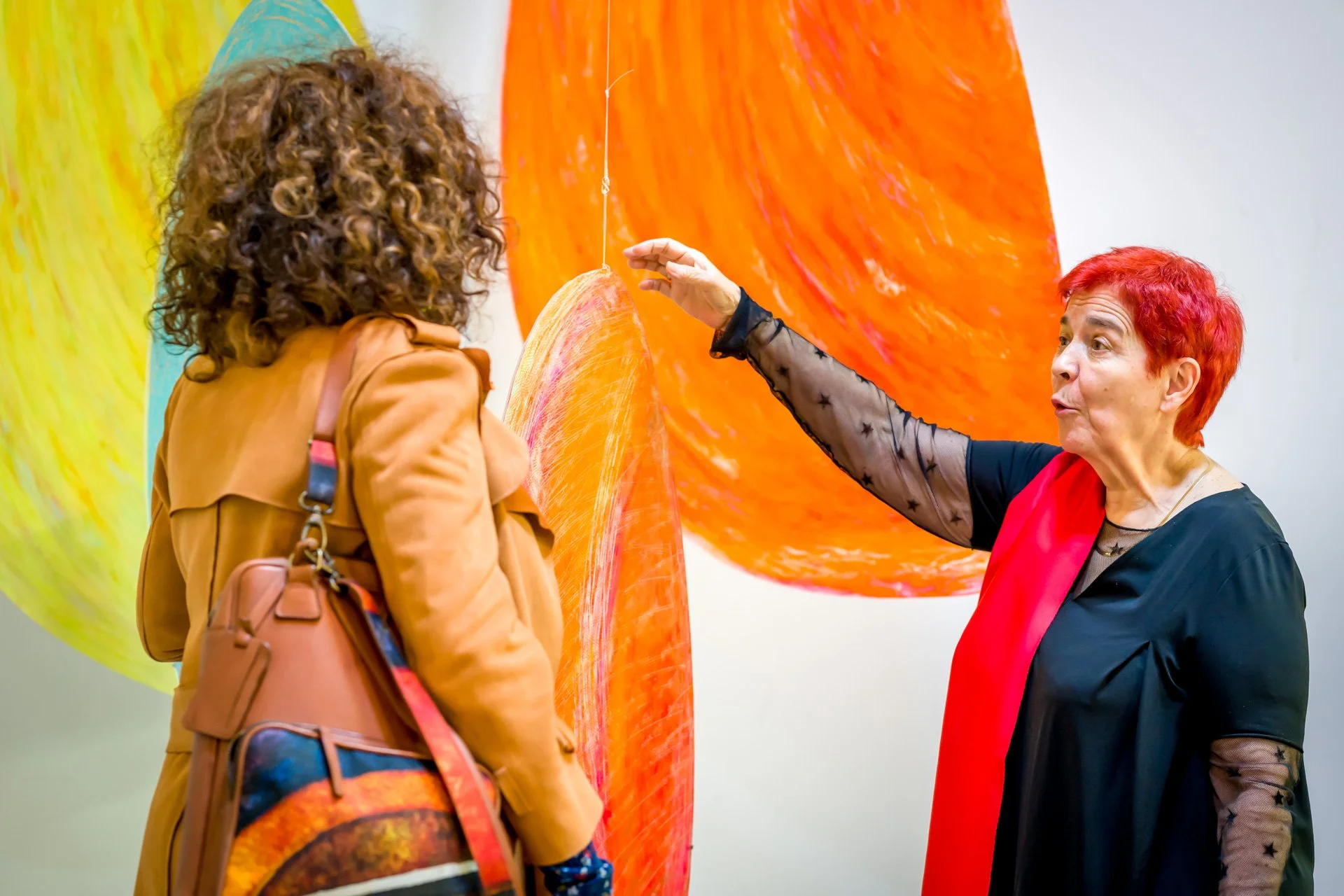 pendant le cocktail de vernissage d'une exposition à Bruxelles. Deux femmes discutent devant des œuvres d'art colorées et suspendues dans une galerie.