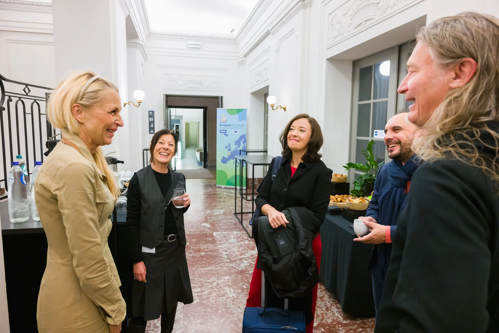 pendant un événement à Bruxelles. Plusieurs personnes souriantes discutent debout dans un espace intérieur élégant, avec des boissons et de la nourriture sur une table en arrière-plan.