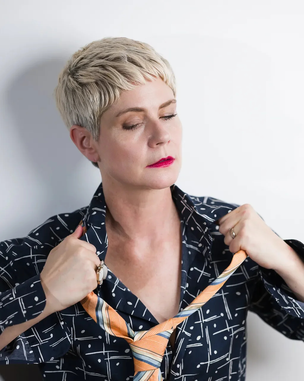 Portrait d’une femme aux cheveux blonds courts, les yeux fermés, ajustant une cravate orange sur une chemise sombre à motifs.