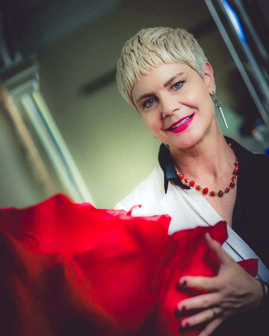 Portrait d’une femme souriante - Stefanie Gasser - aux cheveux blonds courts, portant un collier rouge et tenant un tissu rouge au premier plan.