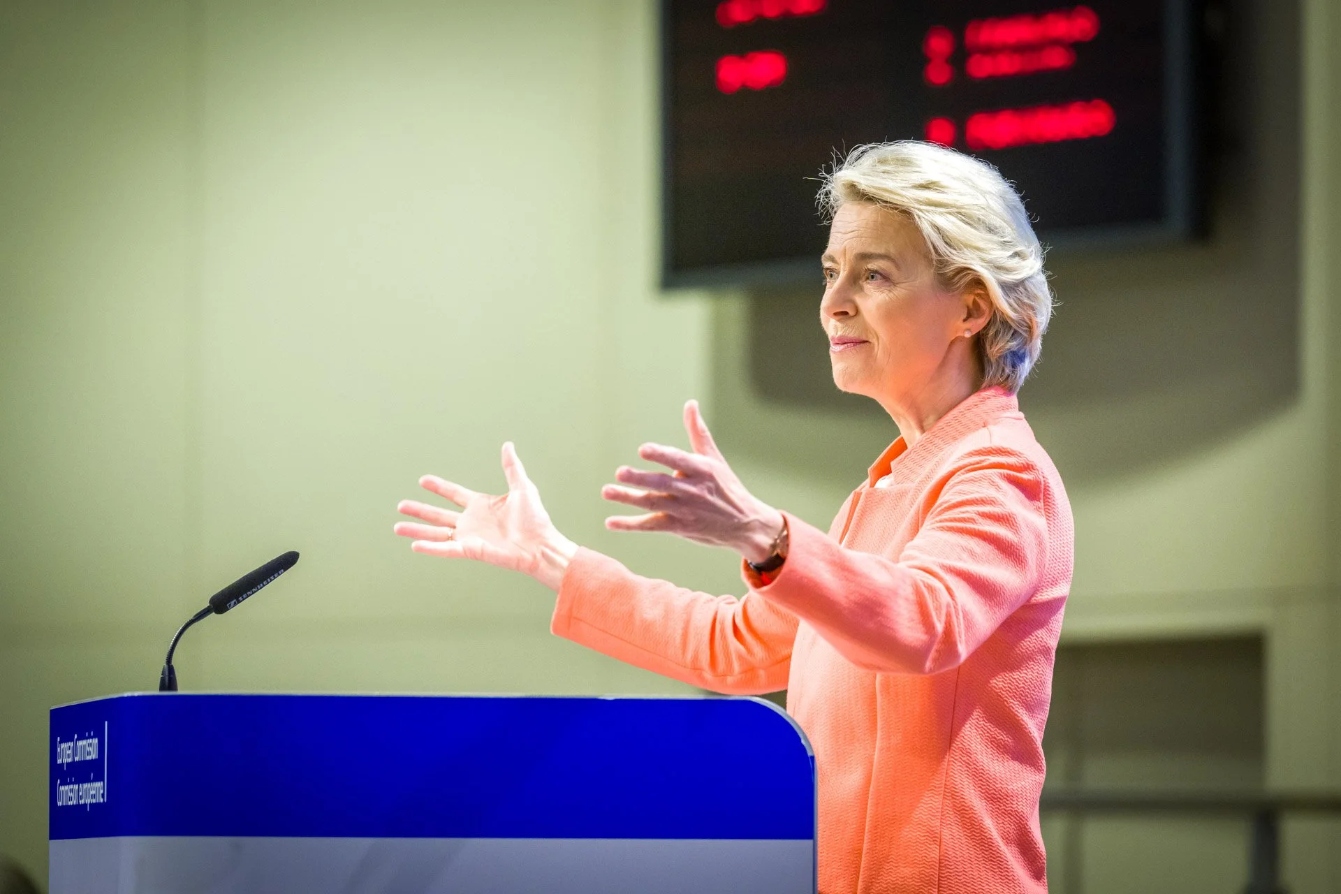 Ursula von der Leyen prise pendant un événement institutionnel à la Commission européenne à Bruxxels. On la voit ici de 3/4 profil, écartant les bras devant un lutriin et en tailleur rose.