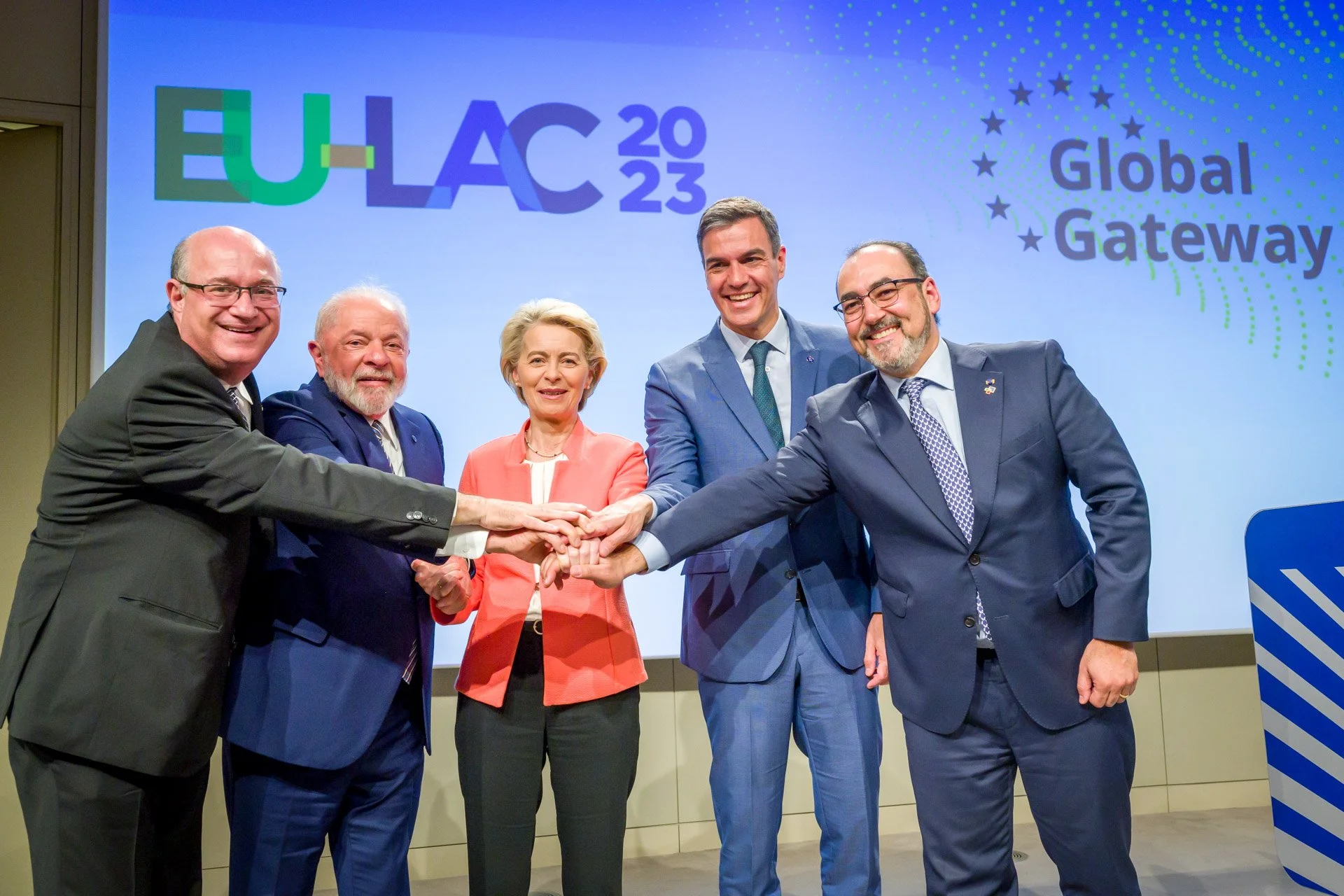 Cinq personnes en costume se tenant la main dans une cérémonie à l'occasion de l'EU-LAC 2023, avec un arrière-plan affichant le texte "EU-LAC 2023" et "Global Gateway".