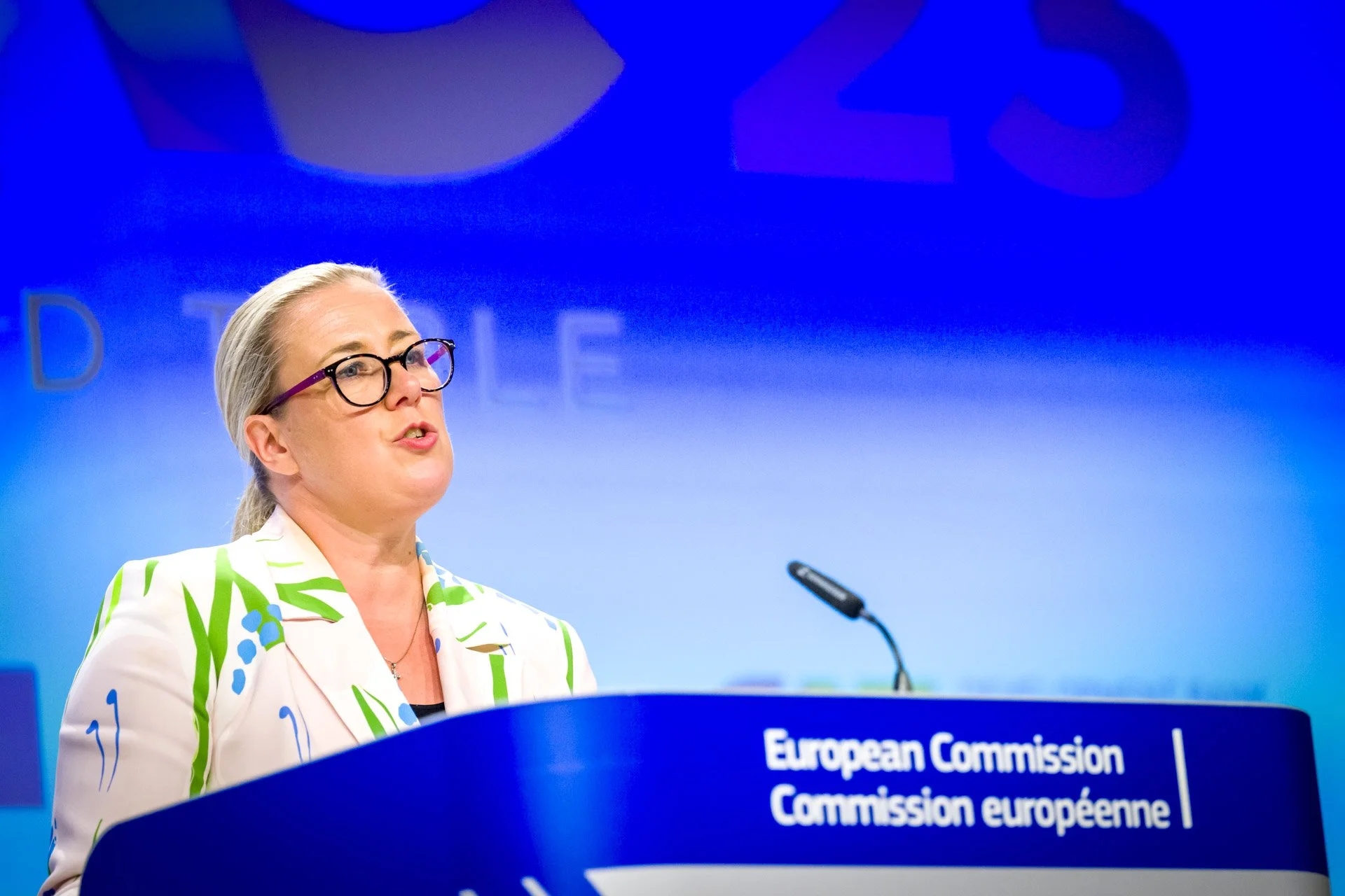 Femme parlant lors d'une conférence de la Commission européenne, portant une veste à motifs verts et bleus, avec un fond bleu et un podium avec le logo de la Commission européenne.