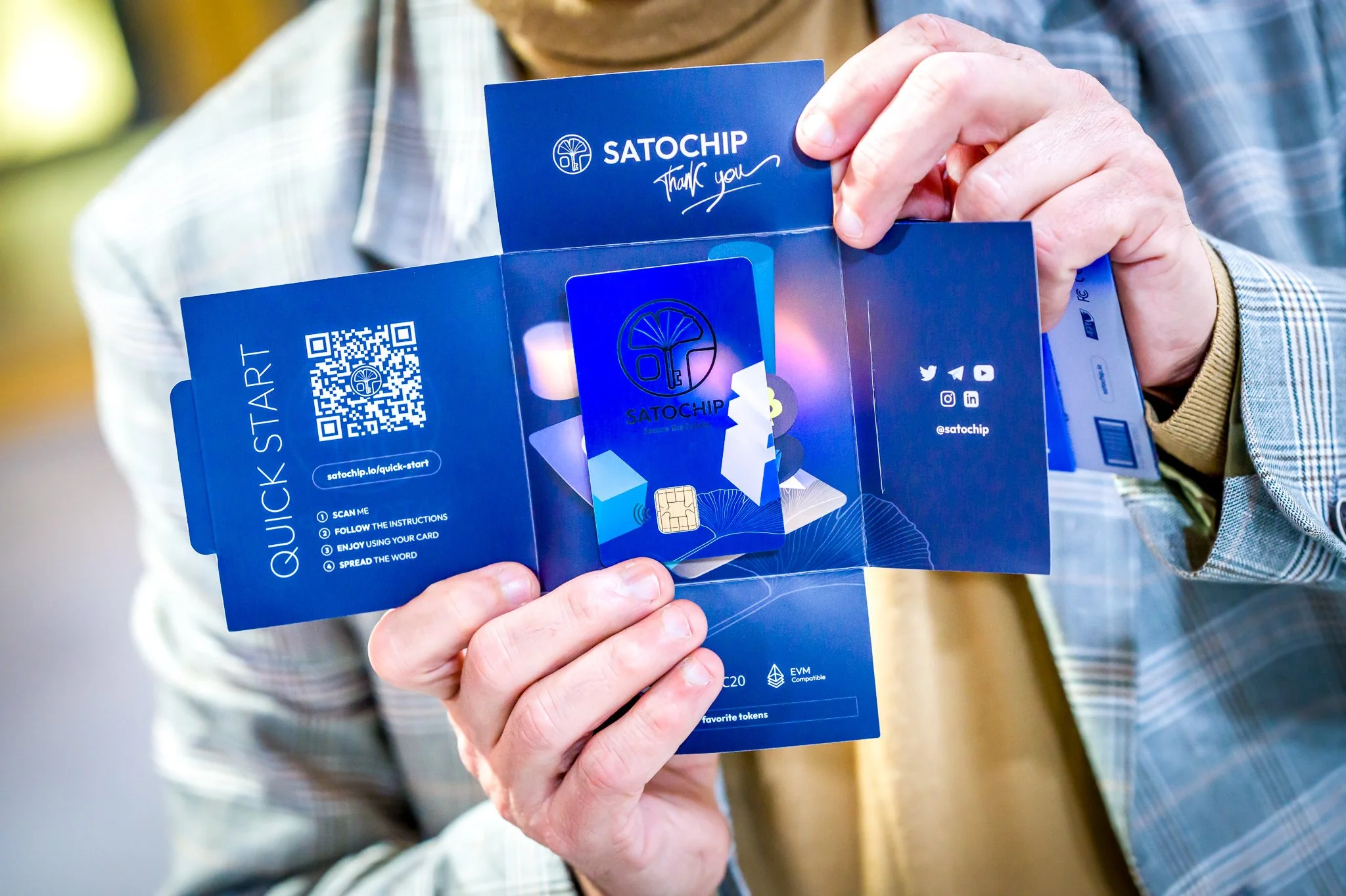 Photographie pendant une conférence internationale à Bruxelles. Personne tenant une brochure bleue avec une carte "Satochip" au centre, QR code et instructions de démarrage rapide sur la gauche, logos des réseaux sociaux à droite.