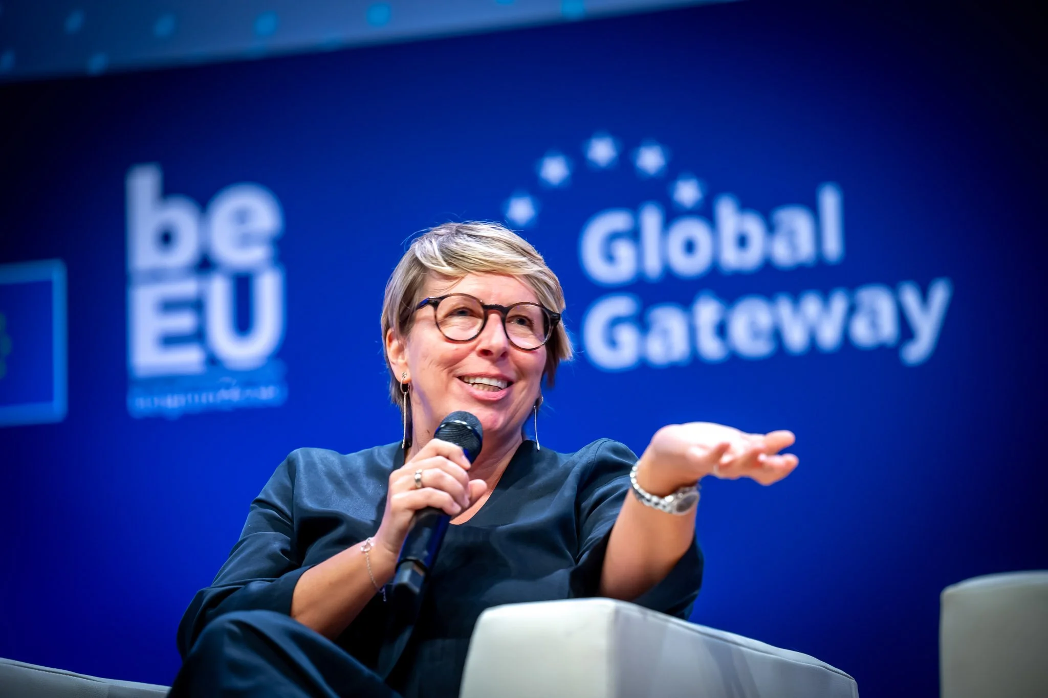 Une ministre belge lors d'un événement international. La ministre tenant un micro avec une toile de fond bleue indiquant "Global Gateway" et "be EU". Elle semble en train de parler lors d'un événement.