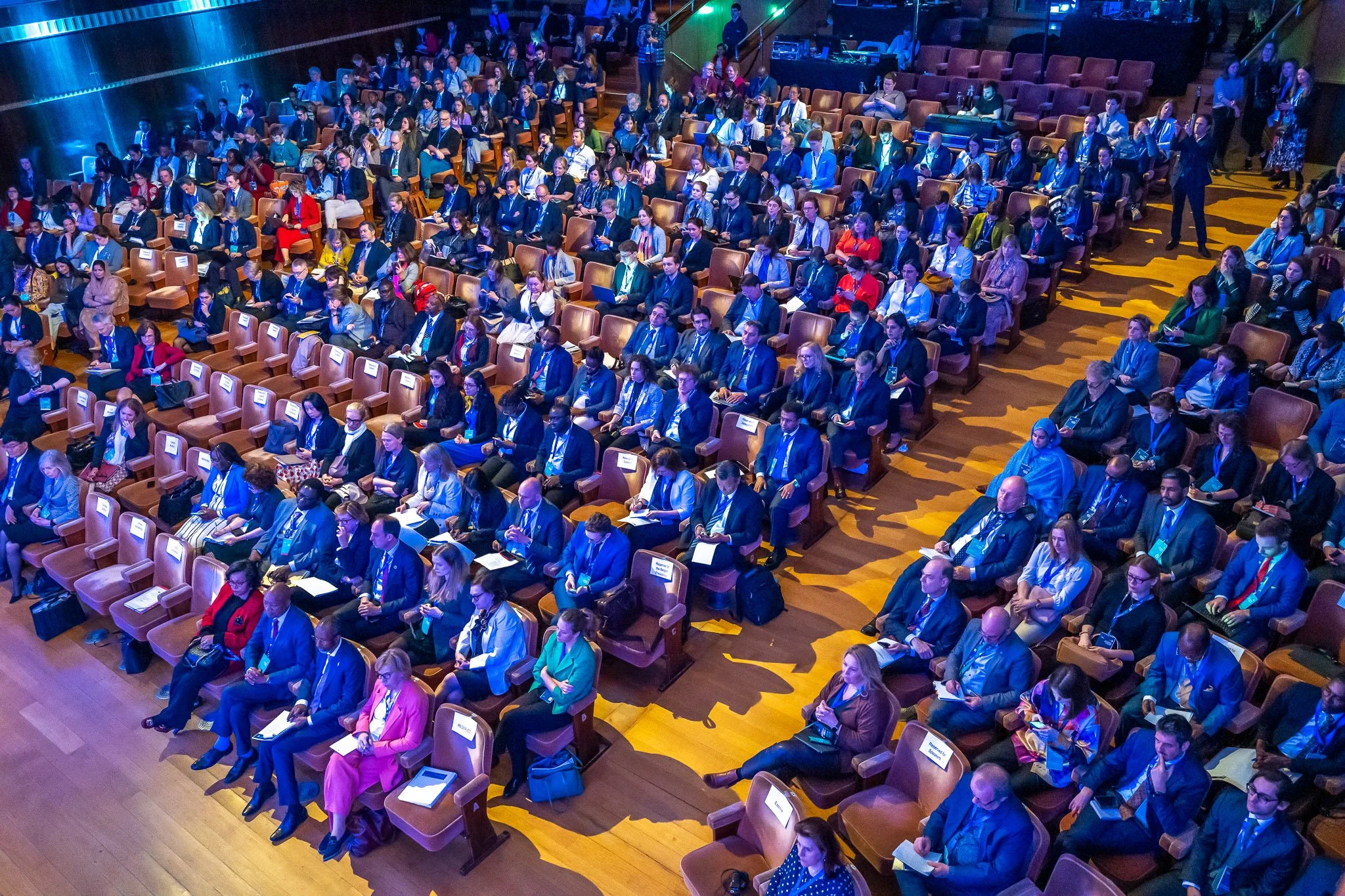 Vue en plongée sur un auditorium rempli de participants assis en rangées, vêtus de costumes, dans une salle éclairée de bleus et d'oranges.